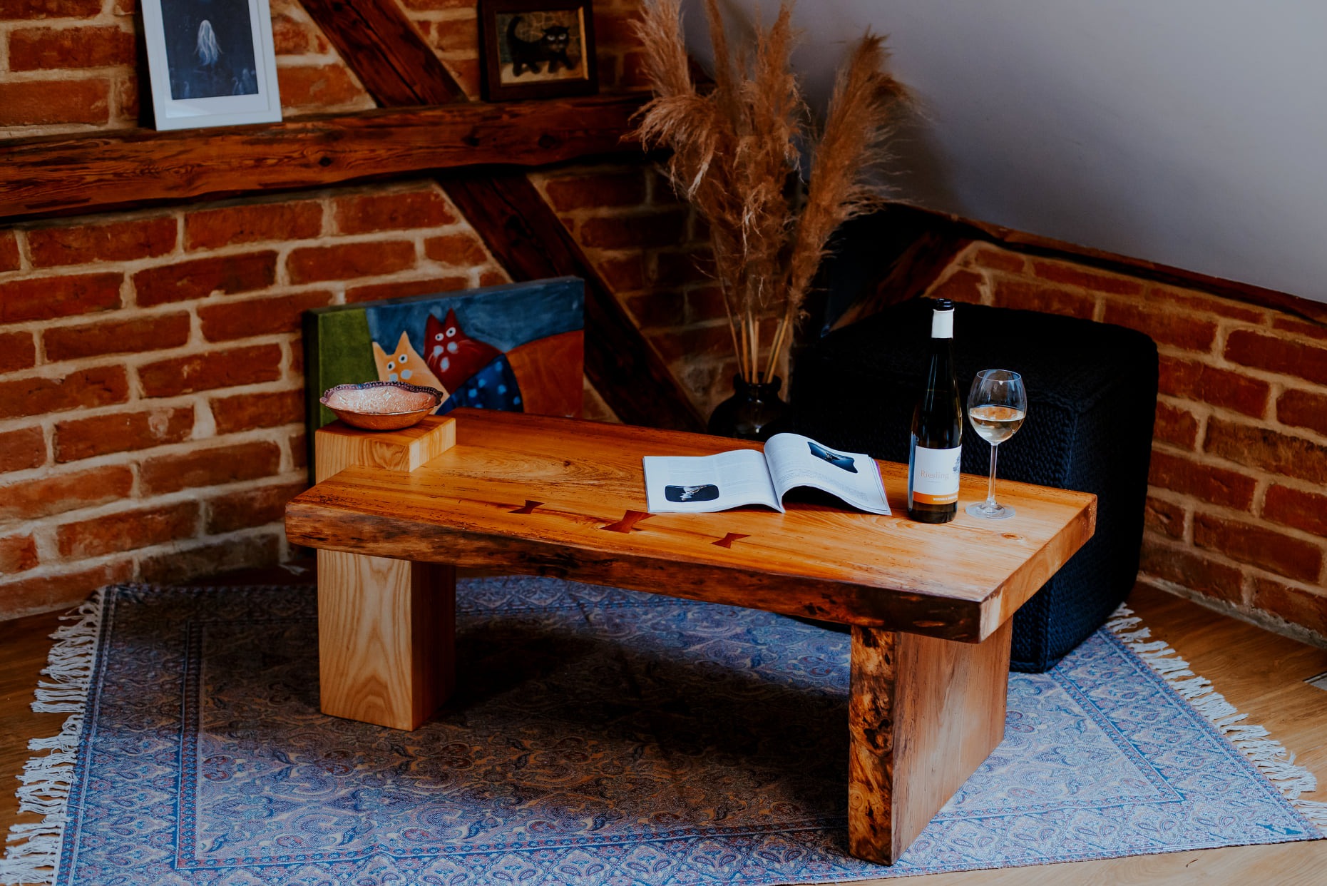 Stolik kawowy z litego drewna z naturalnymi krawędziami, połączony wstawkami w kształcie strzałek, na tle ceglanej ściany i wzorzystego dywanu, z butelką wina i otwartą książką