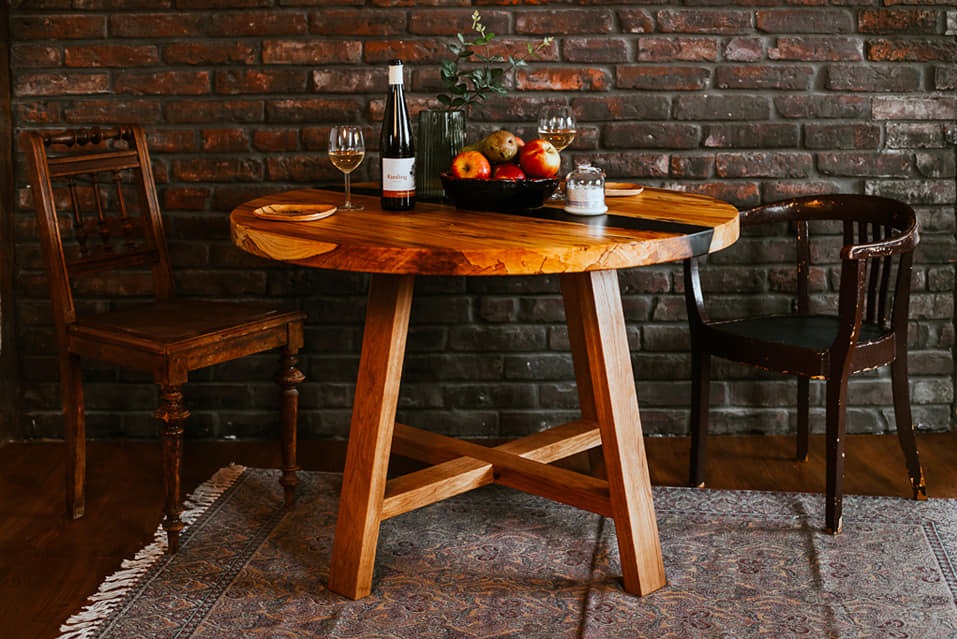 Okrągły stół z blatem z surowego drewna i żywicą epoksydową, ustawiony na drewnianym dywanie, z butelką wina, owocami i dwoma krzesłami w tle ceglanej ściany.