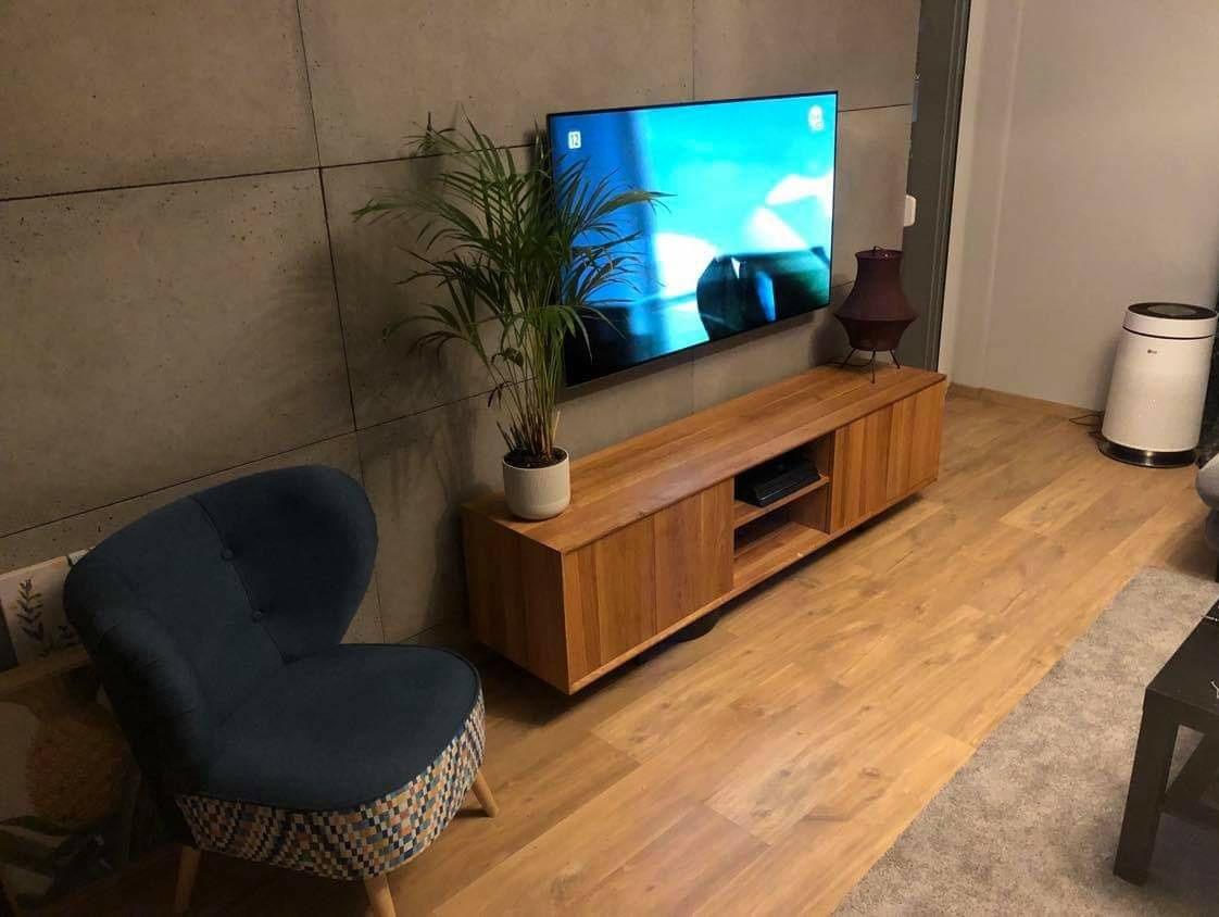 Drewniana szafka RTV z przesuwnymi drzwiczkami, umieszczona pod telewizorem na ścianie z betonowych płyt. Obok stoi fotel z ciemnoniebieskim obiciem i wzorzystym dołem oraz doniczka z rośliną.