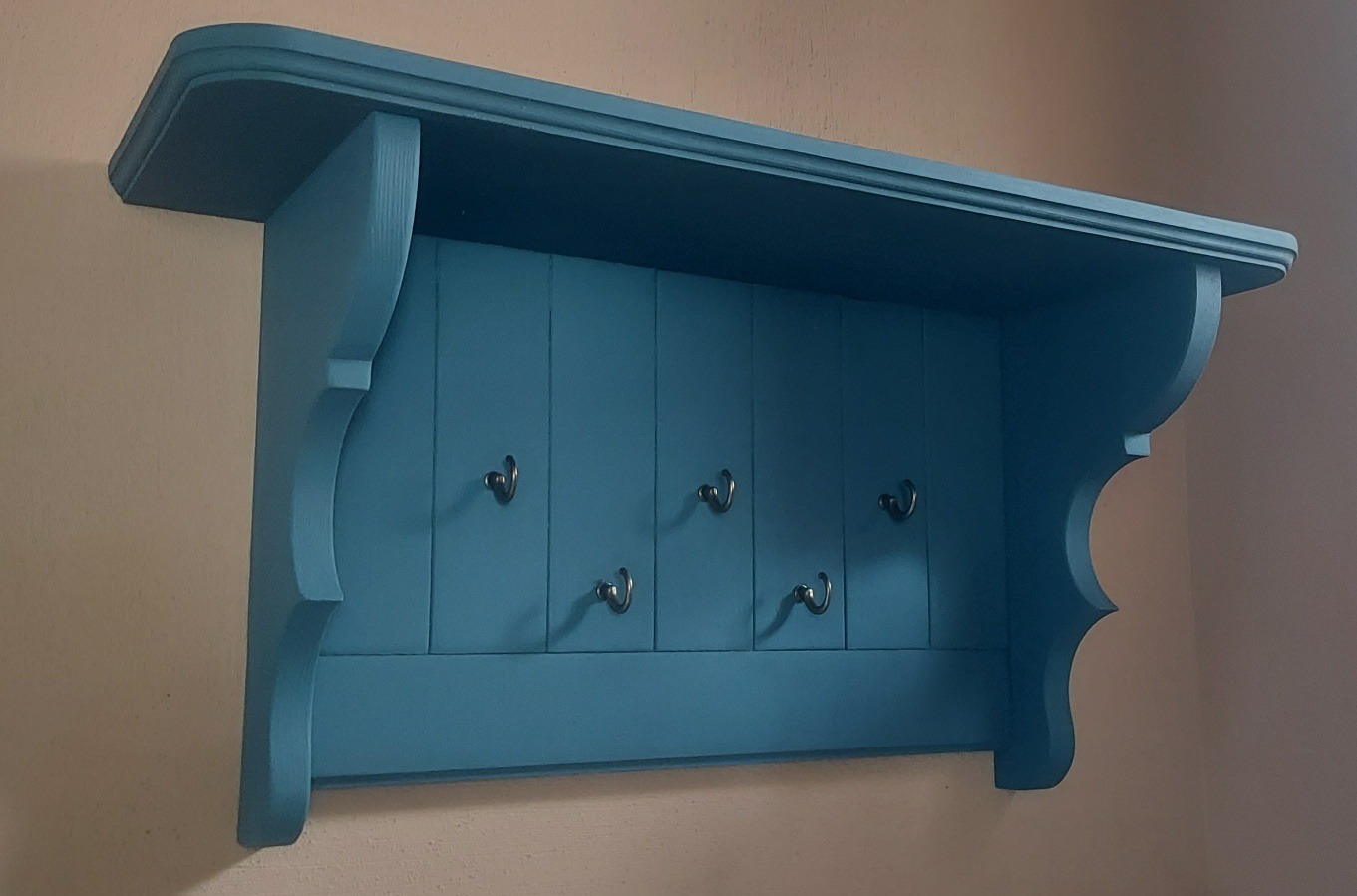 Błękitna, drewniana półka z wieszakami, zawieszona na ścianie, z dekoracyjnymi wykończeniami krawędzi.