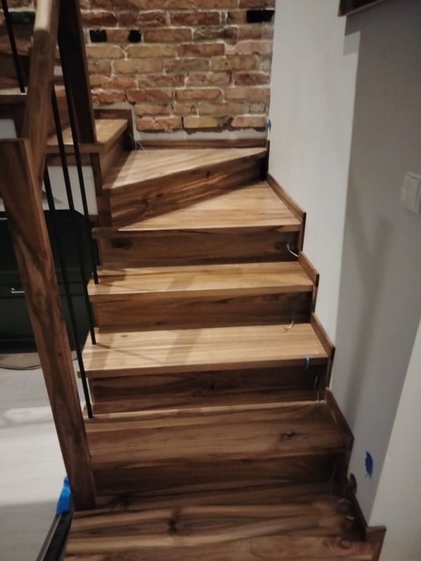 Drewniane schody z wyraźnym usłojeniem, czarne metalowe balustrady, ściana z czerwonej cegły w tle, widoczne niebieskie zabezpieczenia na ścianie.