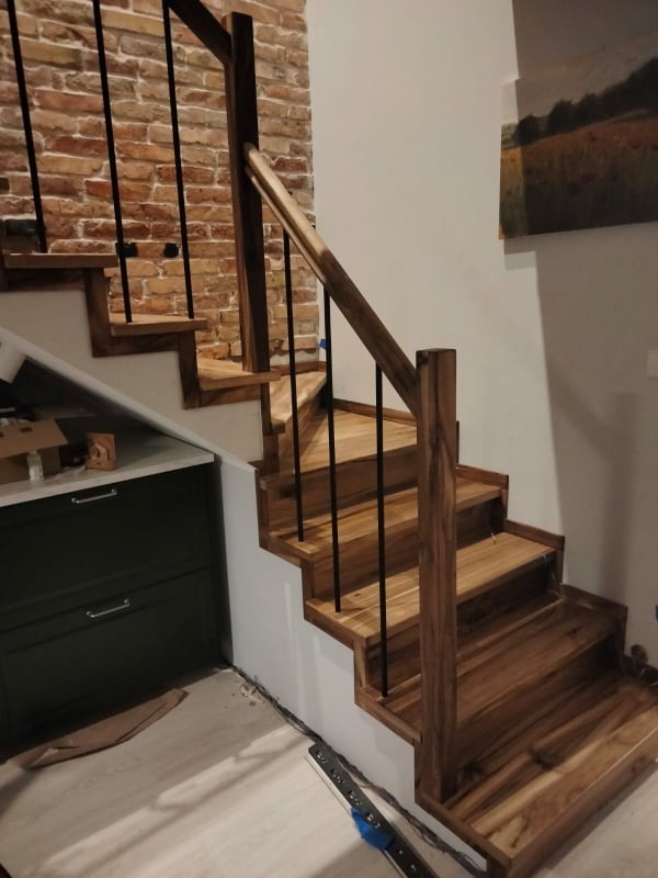 Nowoczesne schody z drewna egzotycznego z czarnymi, metalowymi balustradami, na tle ściany z cegły i obrazu, widoczne niedokończone wykończenie podłogi.
