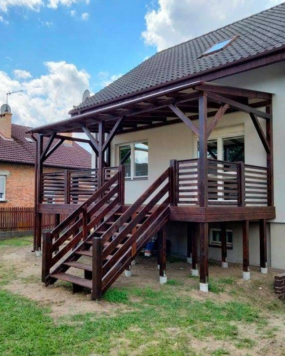 Drewniany taras z zadaszeniem i schodami w Kłodzku, z widoczną konstrukcją wspartą na słupach i balustradą z poziomych desek, prowadzący do domu z białymi ścianami i brązowym dachem.