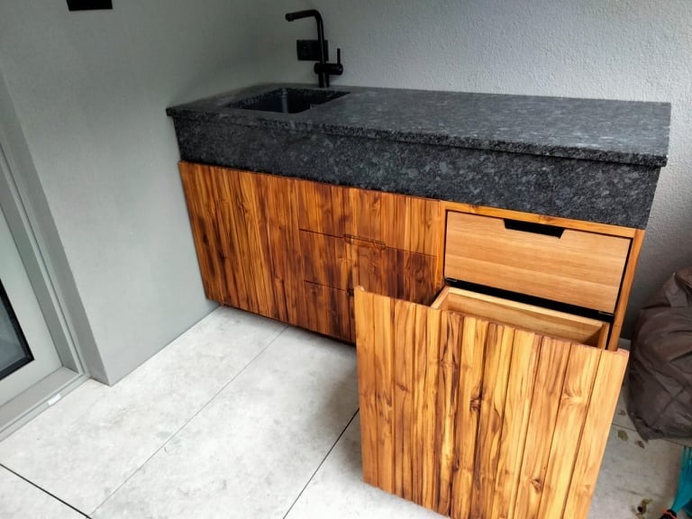 Zewnętrzna szafka z blatem z ciemnego granitu i drewnianymi frontami, jeden front otwarty, widoczny zlew i czarna bateria, szare ściany i jasna podłoga.