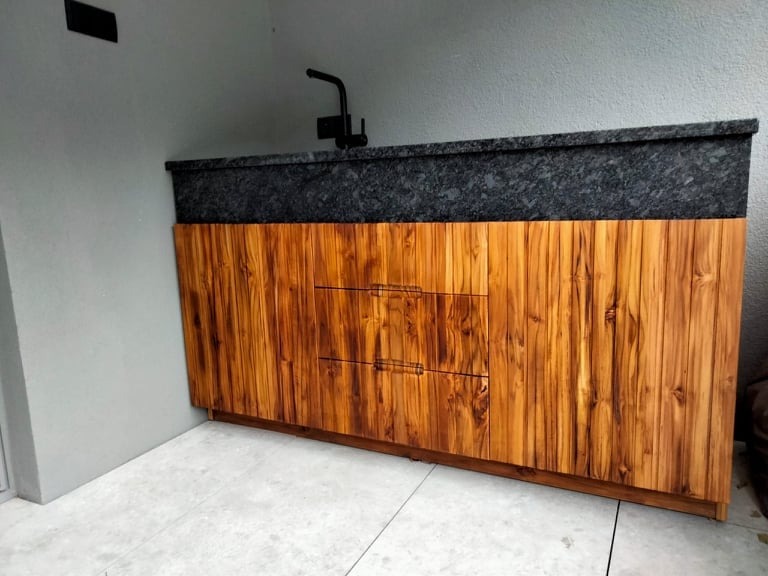 Drewniana szafka z blatem z ciemnego kamienia i czarną armaturą, umieszczona na zewnątrz, prawdopodobnie jako element kuchni letniej.