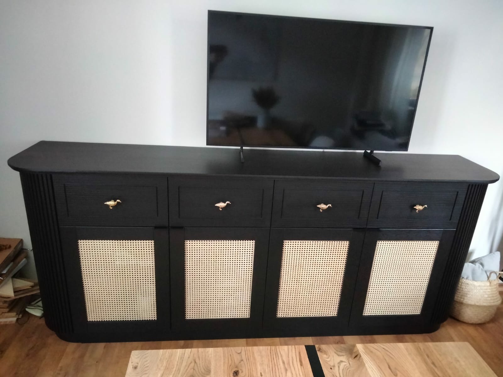Czarna komoda RTV z czterema szufladami i ażurowymi drzwiczkami z rattanu, z uchwytami w kształcie ptaków, stojąca na drewnianej podłodze pod telewizorem.