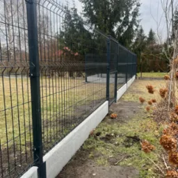 Ciemny panel ogrodzeniowy na betonowej podmur&oacute;wce, widok z boku, trawa i drzewa w tle, pochmurne niebo. Nowoczesne ogrodzenie działki.