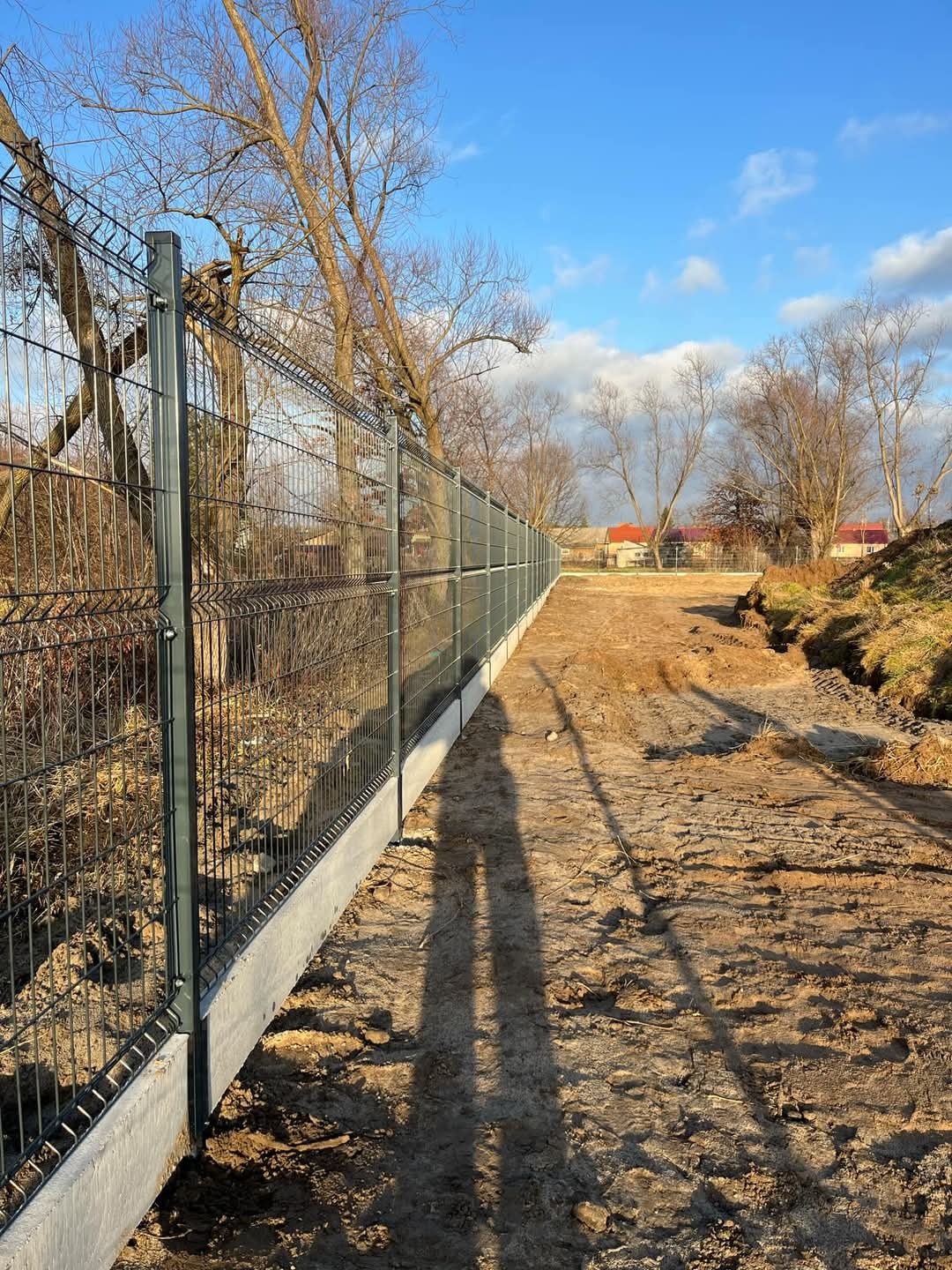 Nowoczesne ogrodzenie panelowe z podmurówką betonową, widoczne z perspektywy ziemi, z długim cieniem osoby fotografującej na pierwszym planie.