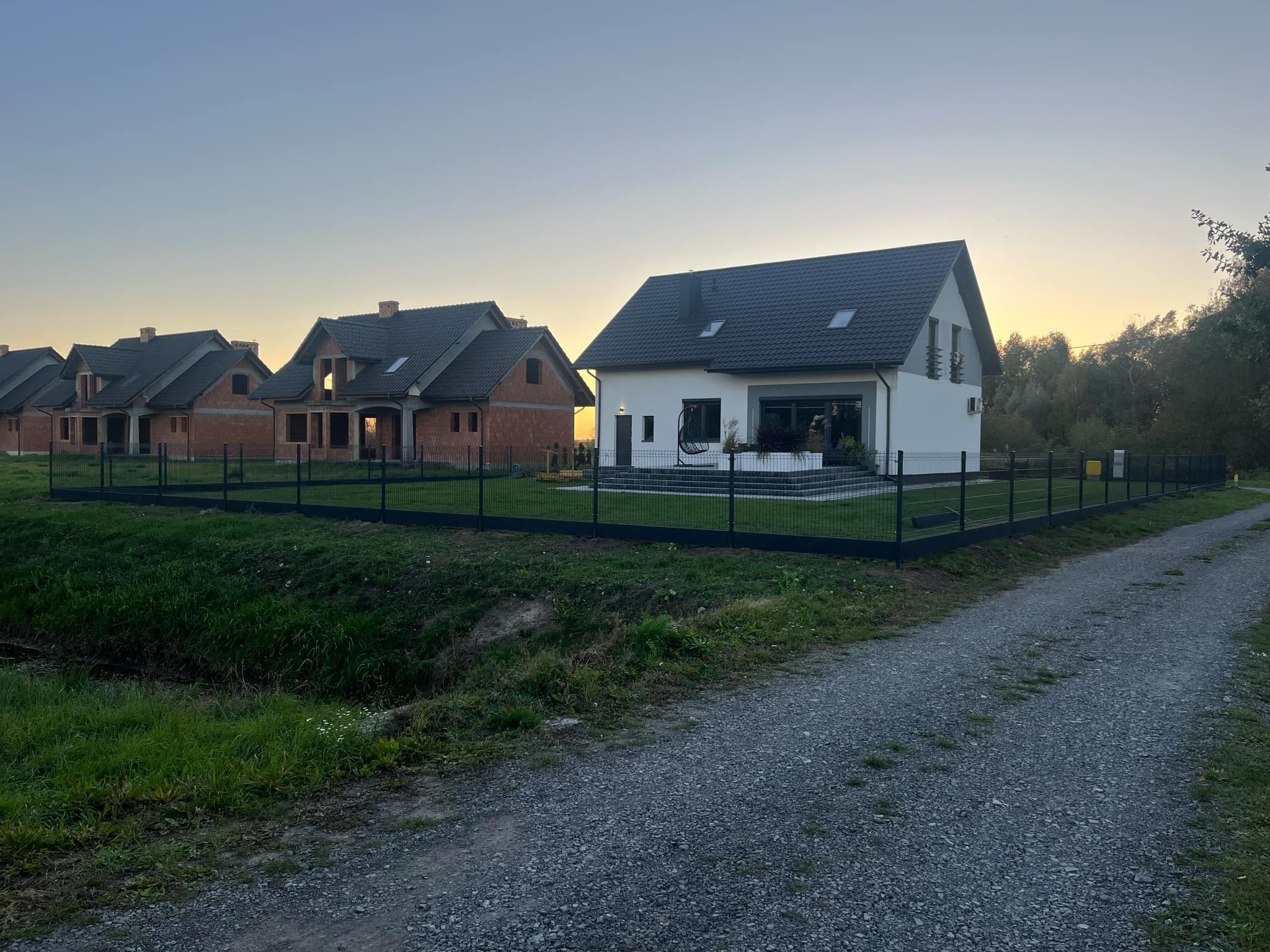 Nowoczesny dom jednorodzinny z czarnym ogrodzeniem panelowym w Tarnobrzegu. W tle domy w stanie surowym i zachodzące słońce.