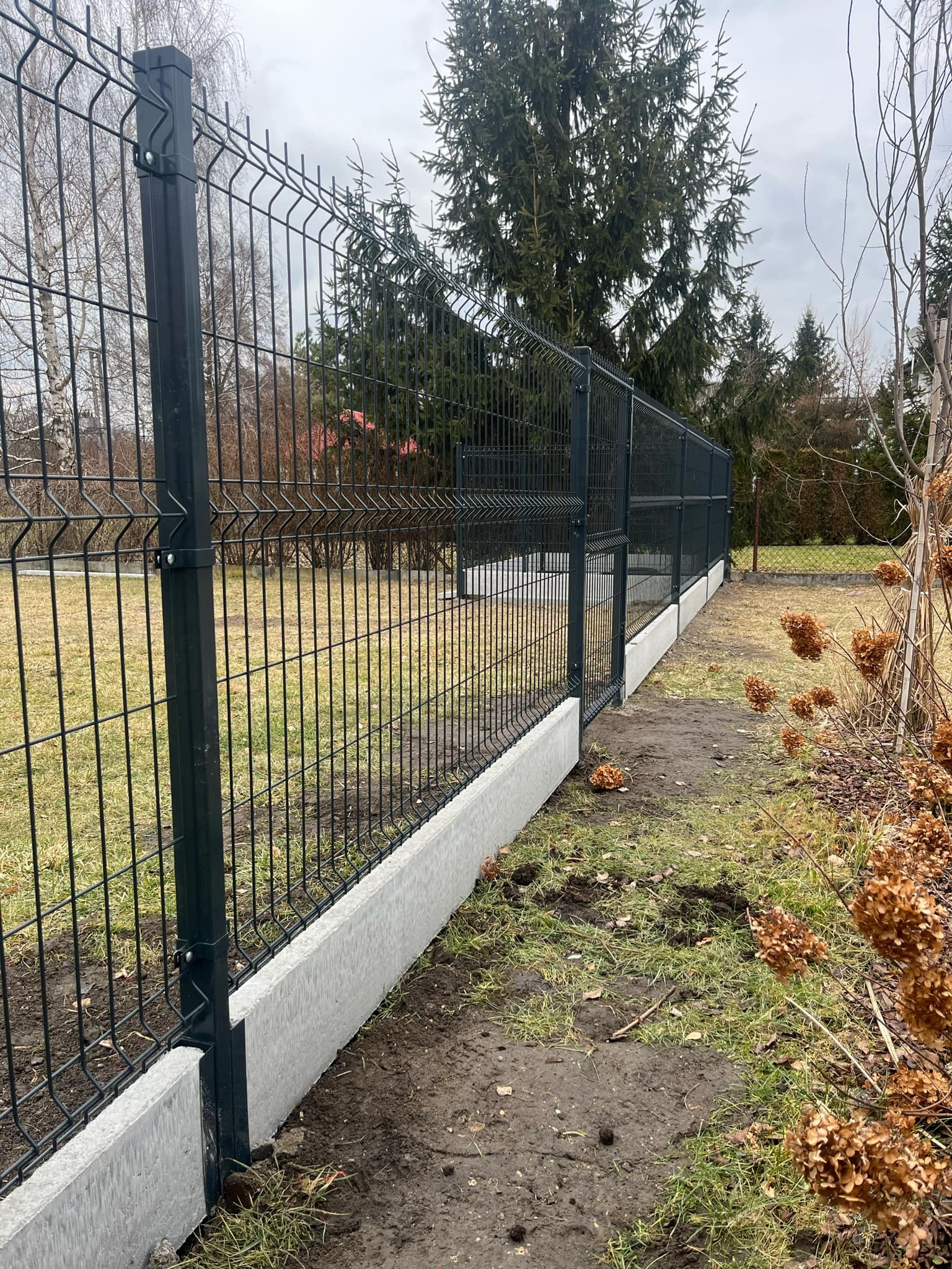 Ciemny panel ogrodzeniowy na betonowej podmurówce, widok z boku, trawa i drzewa w tle, pochmurne niebo. Nowoczesne ogrodzenie działki.