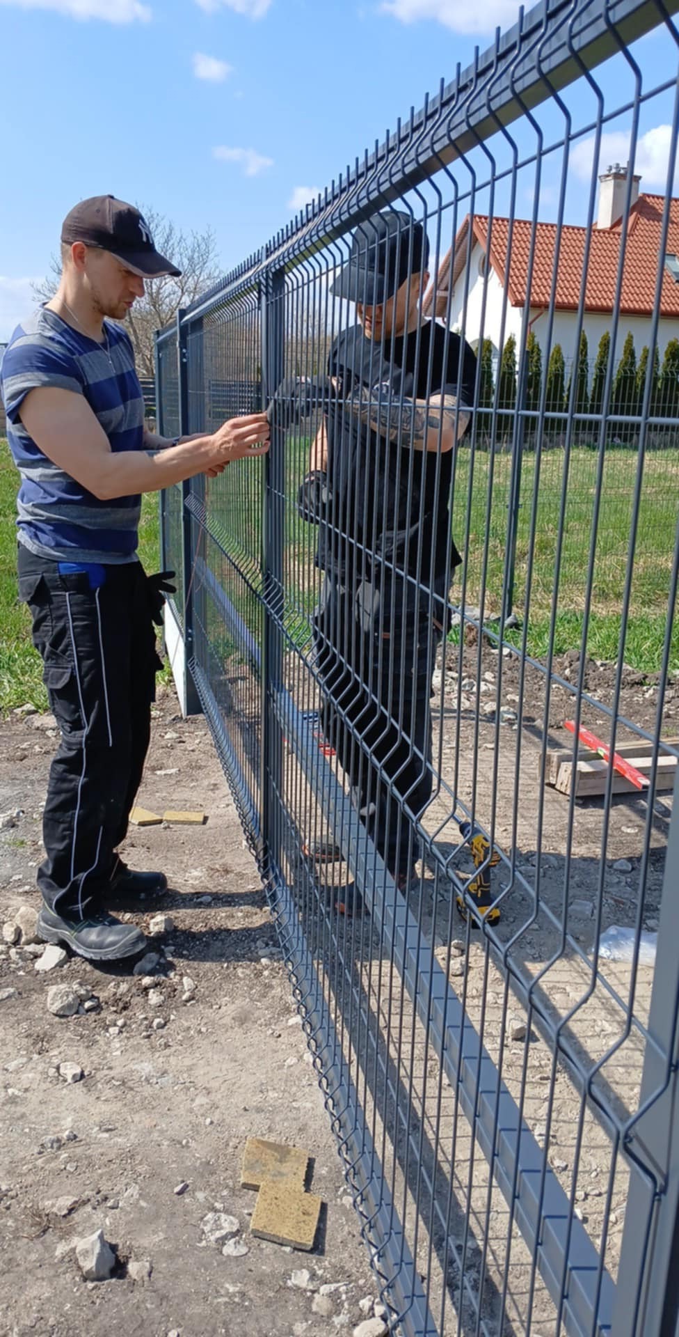 Dwóch monterów łączy elementy szarego ogrodzenia panelowego na tle domu z czerwoną dachówką i błękitnego nieba. Widoczne narzędzia i fragment gruntu.
