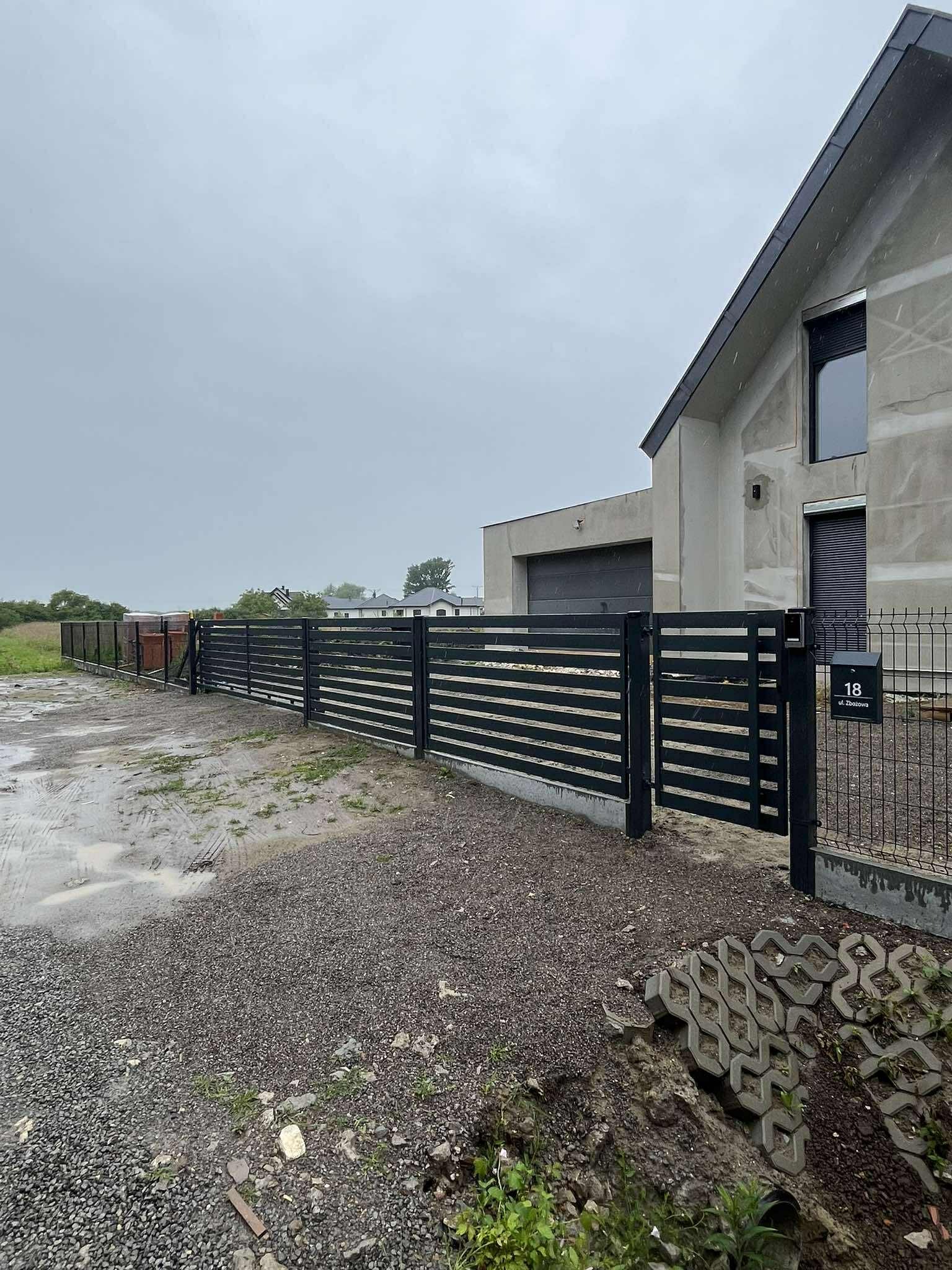 Nowoczesne, poziome ogrodzenie panelowe w kolorze czarnym, z bramką i skrzynką pocztową na tle surowego domu w trakcie budowy. Widok na posesję w pochmurny dzień.