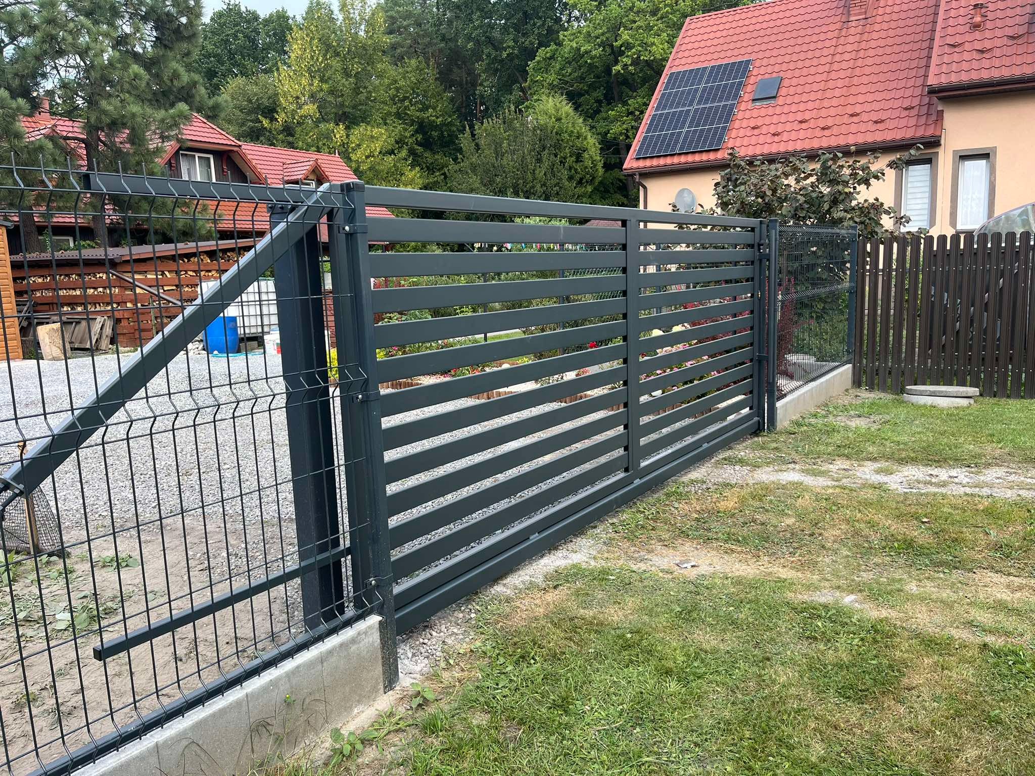 Nowoczesna, pozioma brama przesuwna z szarego metalu, zintegrowana z ogrodzeniem panelowym. Widok na dom z czerwonym dachem i panele słoneczne w tle.