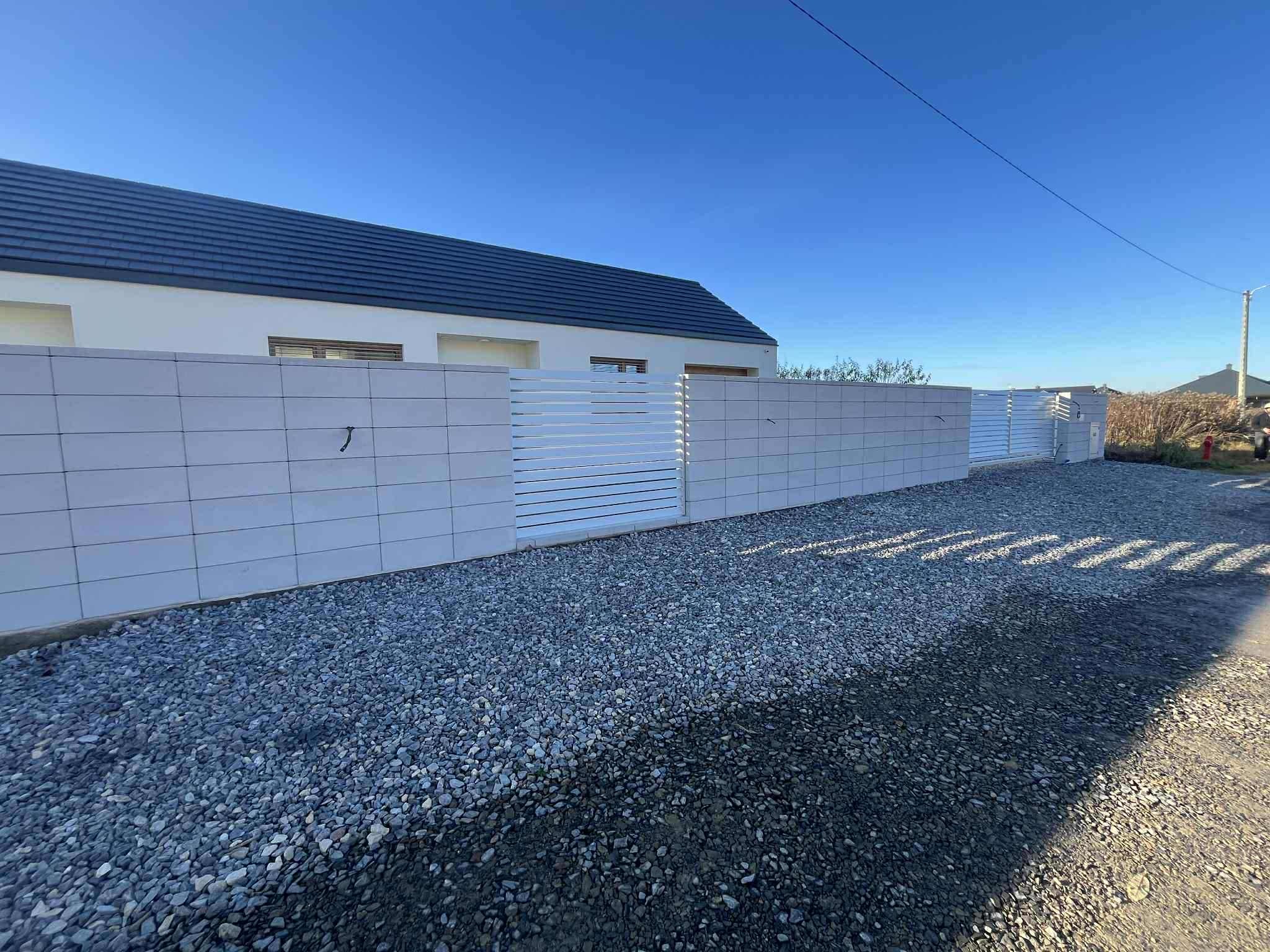 Nowoczesne ogrodzenie z białych, betonowych bloczków i poziomych, białych paneli, z bramą wjazdową. Podłoże wysypane szarym żwirem, w tle dom z ciemnym dachem.