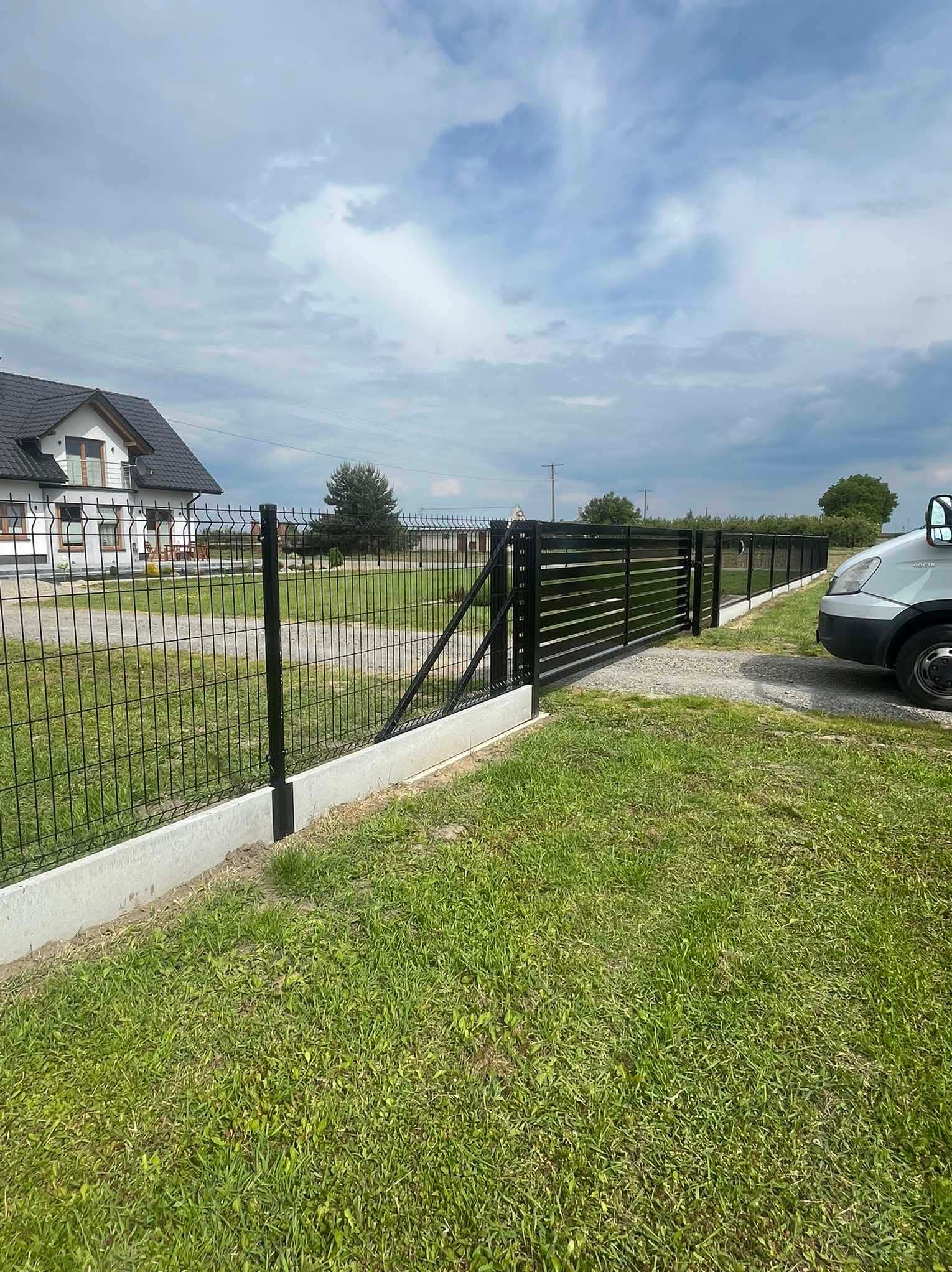Nowoczesne ogrodzenie panelowe z bramą przesuwną, czarne słupki, podmurówka betonowa, widok z zewnątrz, trawnik, dom w tle, częściowo widoczny samochód dostawczy.