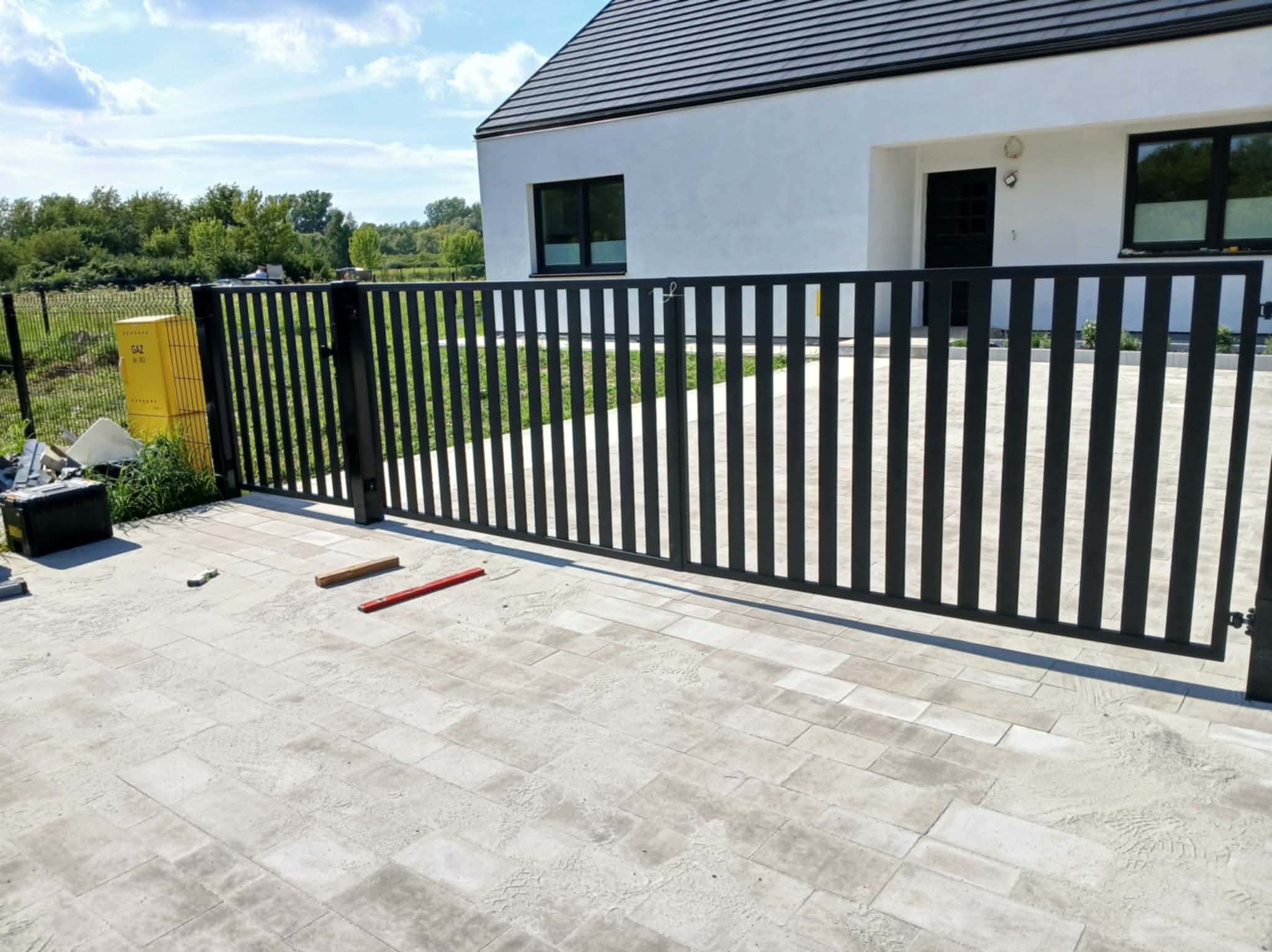 Nowoczesna, czarna brama metalowa z pionowymi szczeblami, zamontowana przed białym domem z ciemną dachówką. Widoczne narzędzia i kostka brukowa.