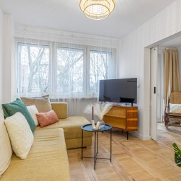 Gradiam Nieruchomości - Jasny salon z żółtą sofą, telewizorem na komodzie w stylu retro i widokiem na sypialnię z wiklinowym fotelem bujanym