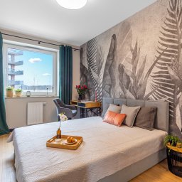 Gradiam Nieruchomości - Jasna sypialnia z łóżkiem, stolikiem nocnym, biurkiem, oknem z widokiem na budynek i dekoracyjną tapetą z motywem liści.