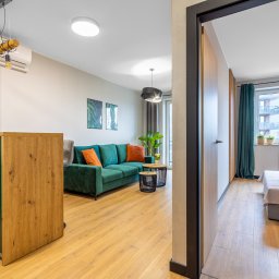 Gradiam Nieruchomości - Jasne, nowocześnie urządzone wnętrze mieszkania z zieloną sofą, drewnianymi akcentami i widokiem na pokój z łóżkiem przez otwarte drzwi.