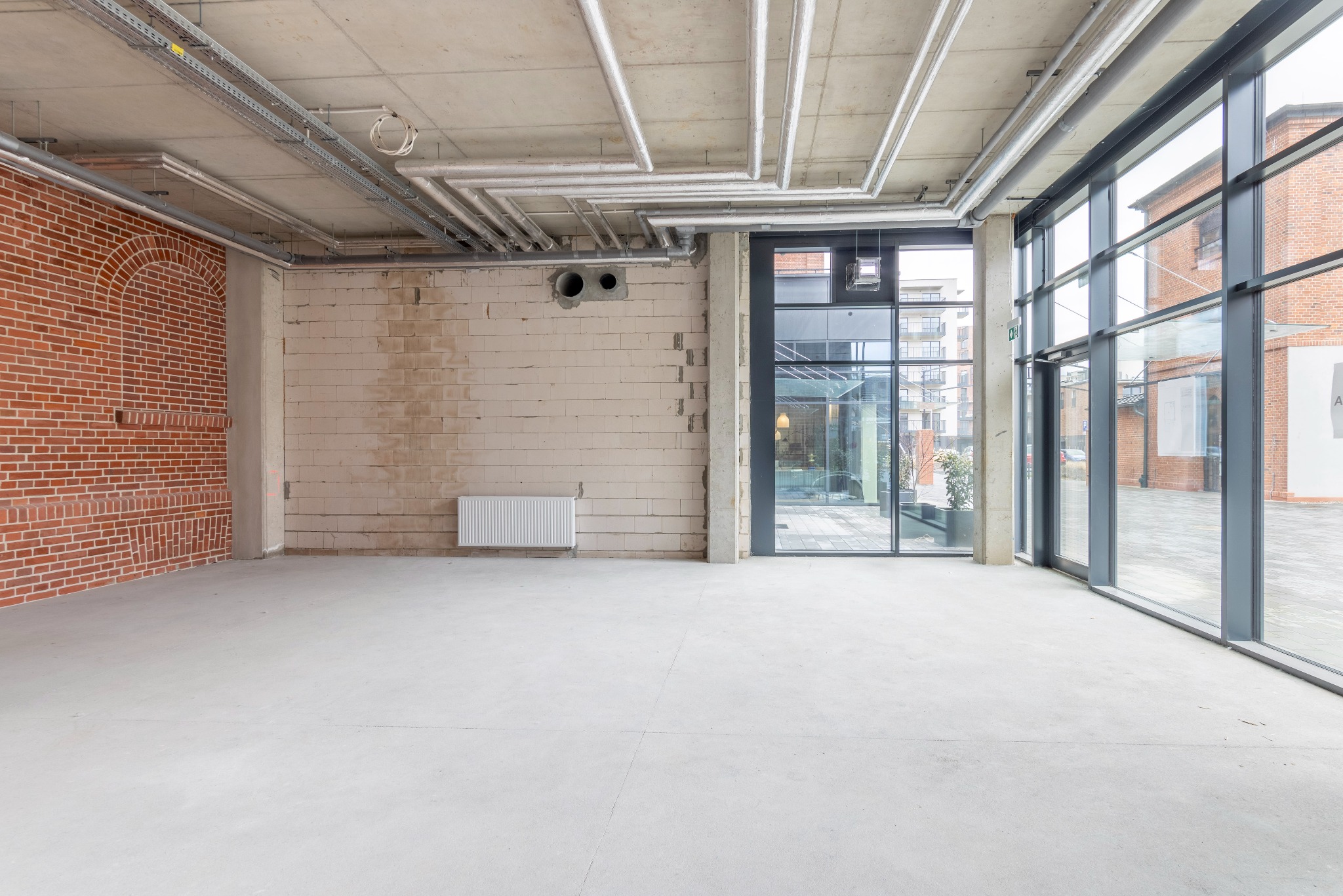 Surowe wnętrze lokalu z widocznymi instalacjami pod sufitem, ceglaną ścianą i dużymi oknami.