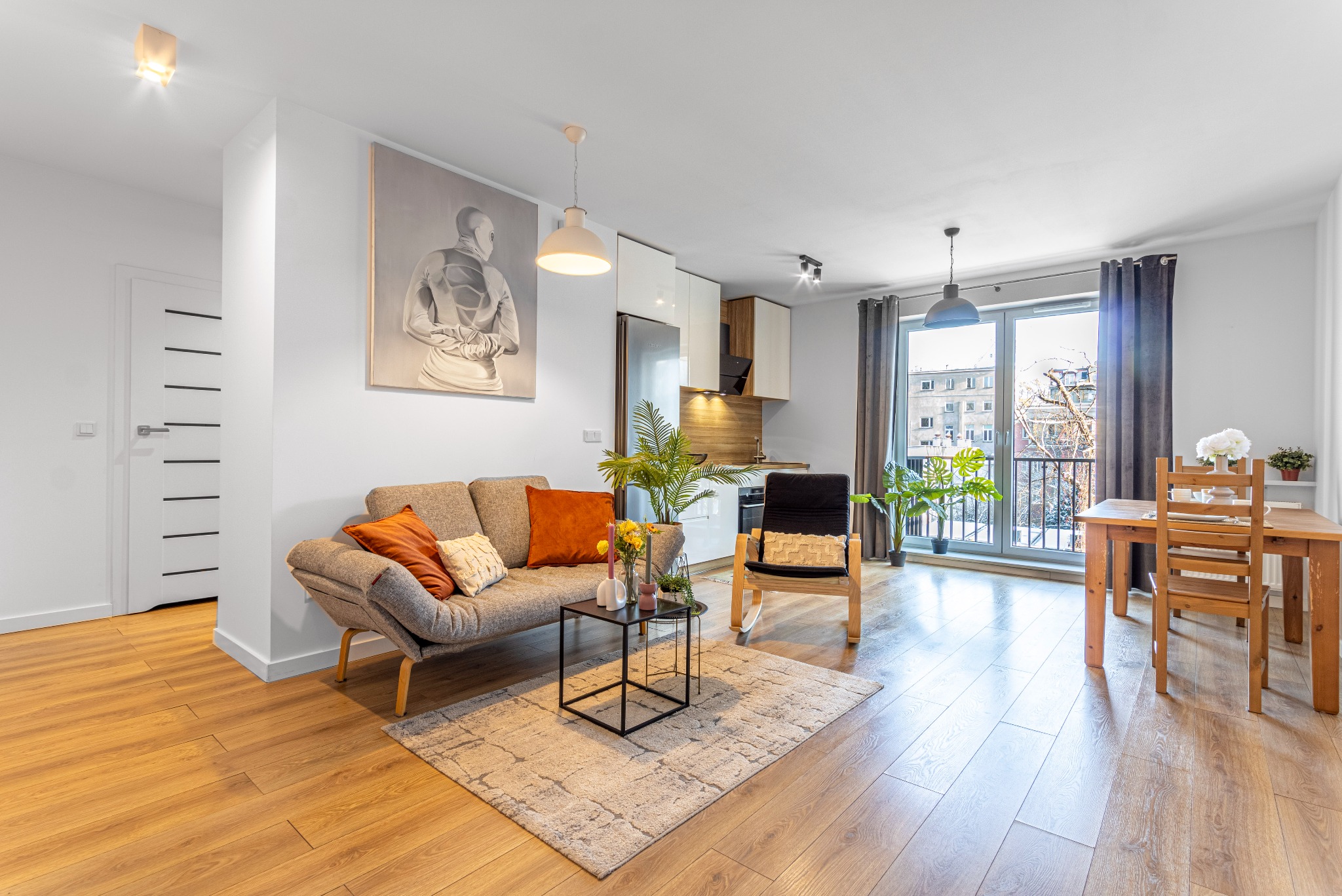 Jasne, przestronne wnętrze mieszkania z drewnianą podłogą, szarą sofą z pomarańczowymi poduszkami, czarnym stolikiem kawowym, drewnianym krzesłem bujanym i stołem jadalnym przy oknie balkonowym...