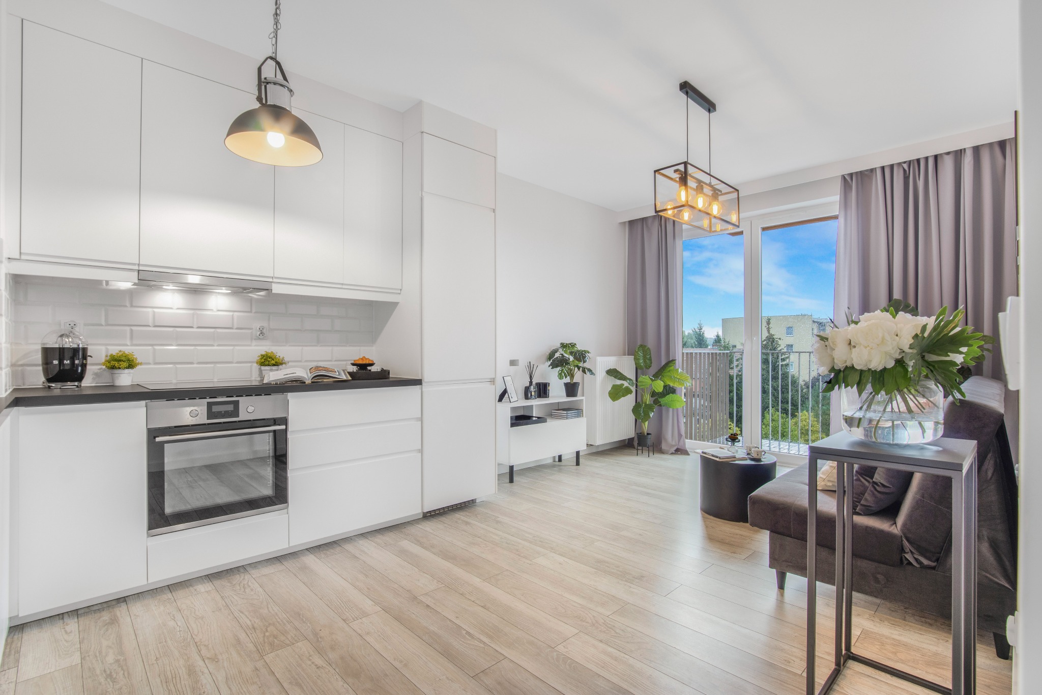 Jasne, nowoczesne wnętrze mieszkania z aneksem kuchennym, drewnianą podłogą i balkonem z widokiem na zieleń, oświetlone lampami wiszącymi.