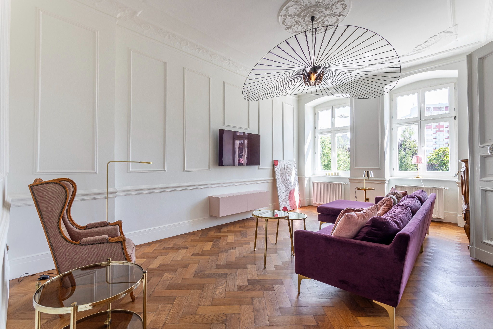 Jasne wnętrze salonu w stylu klasycznym z fioletową sofą, parkietem w jodełkę, białymi ścianami z dekoracyjnymi panelami, designerską lampą i dwoma oknami z widokiem na zieleń.