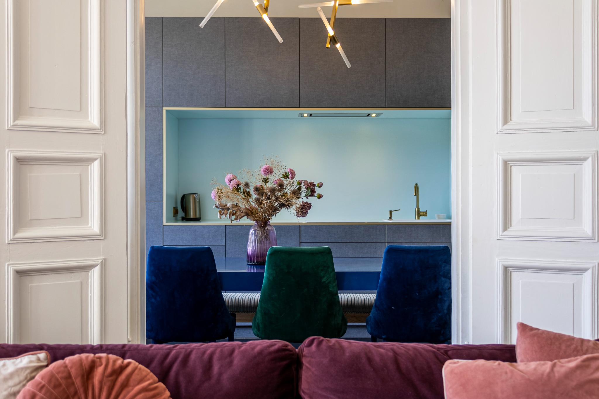 Nowoczesna kuchnia z jadalnią widziana przez otwarte, białe drzwi. W tle szare szafki, turkusowy panel nad blatem, złota armatura i fioletowy wazon z suszonymi kwiatami na stole otoczonym...