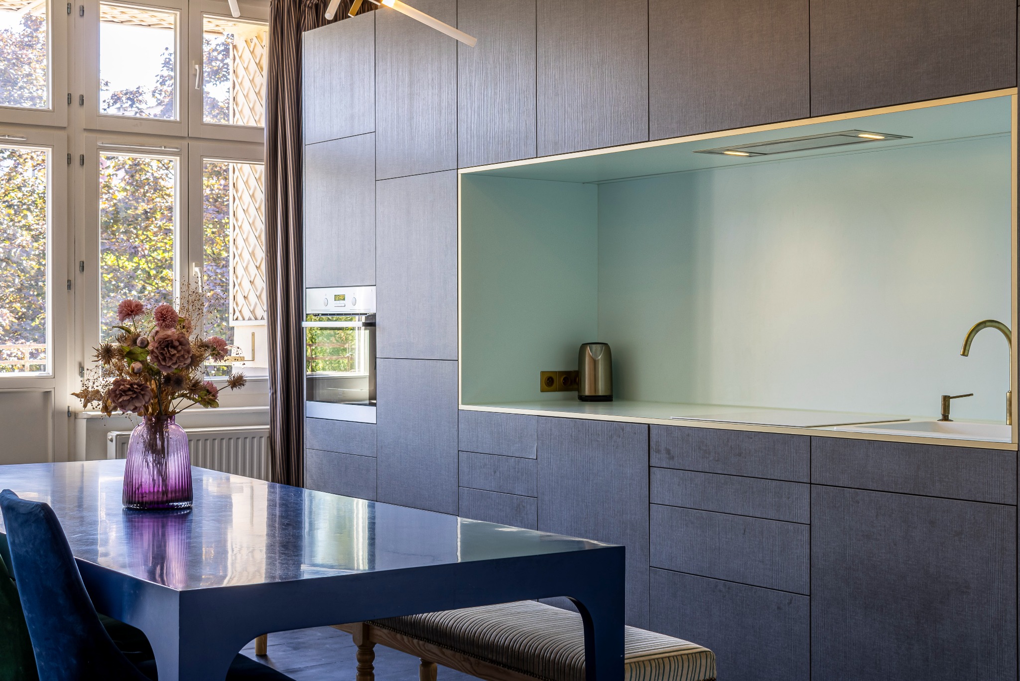 Nowoczesna kuchnia z ciemnoszarymi szafkami, złotymi akcentami i niebieskim stołem z bukietem kwiatów w fioletowym wazonie, oświetlona naturalnym światłem z okna.