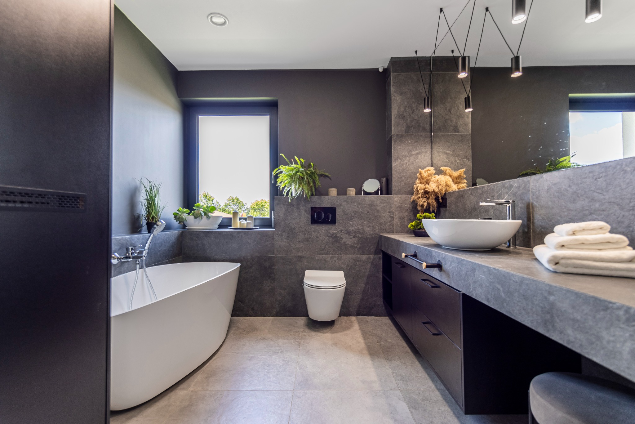 Nowoczesna łazienka w odcieniach szarości z wolnostojącą białą wanną, umywalką nablatową i ciemnymi szafkami podblatowymi, oświetlona wiszącymi lampami punktowymi.