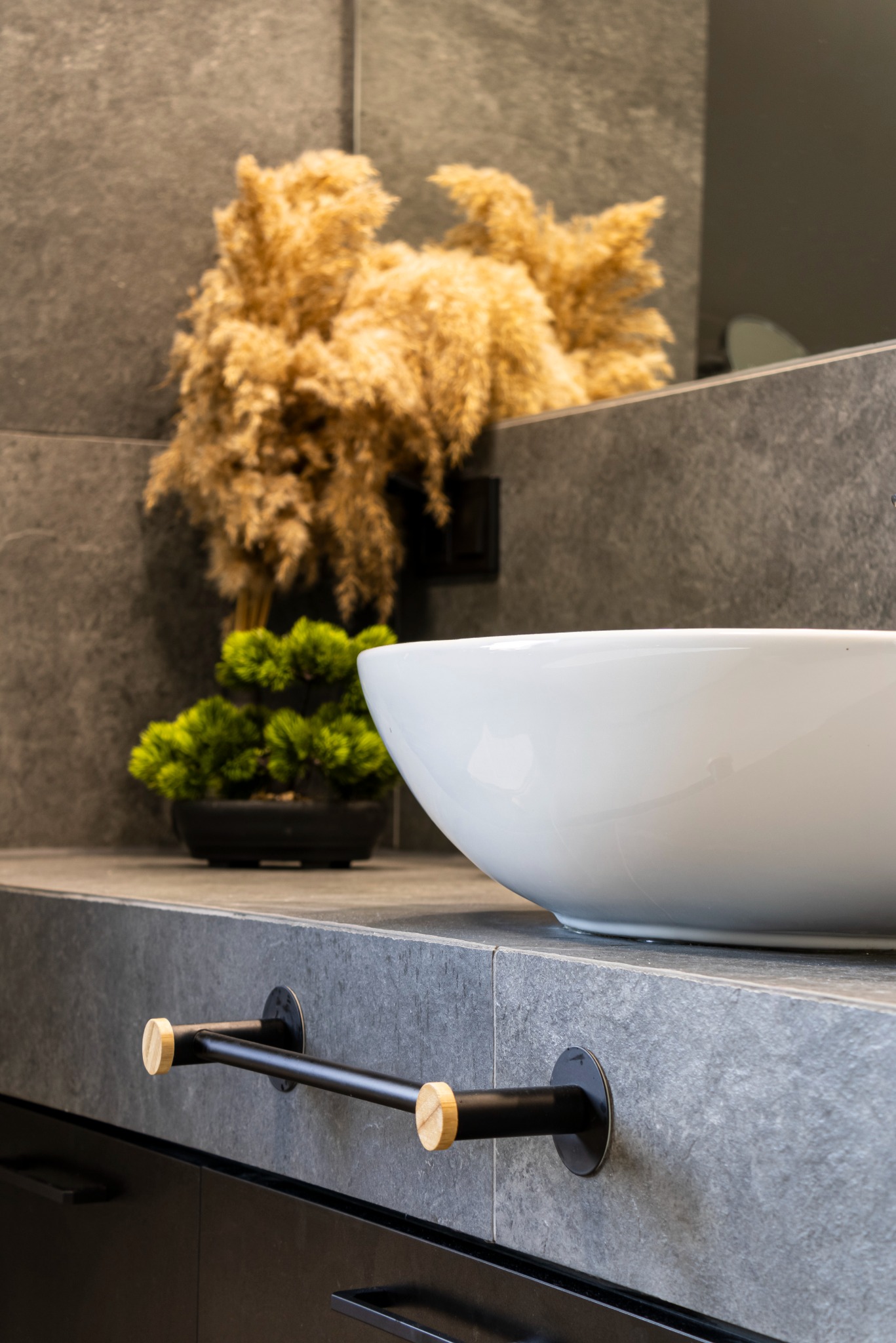 Nowoczesna łazienka z umywalką nablatową, szarą kamienną płytą i czarnym uchwytem na ręczniki z drewnianymi akcentami oraz dekoracyjną trawą pampasową i bonsai w tle.