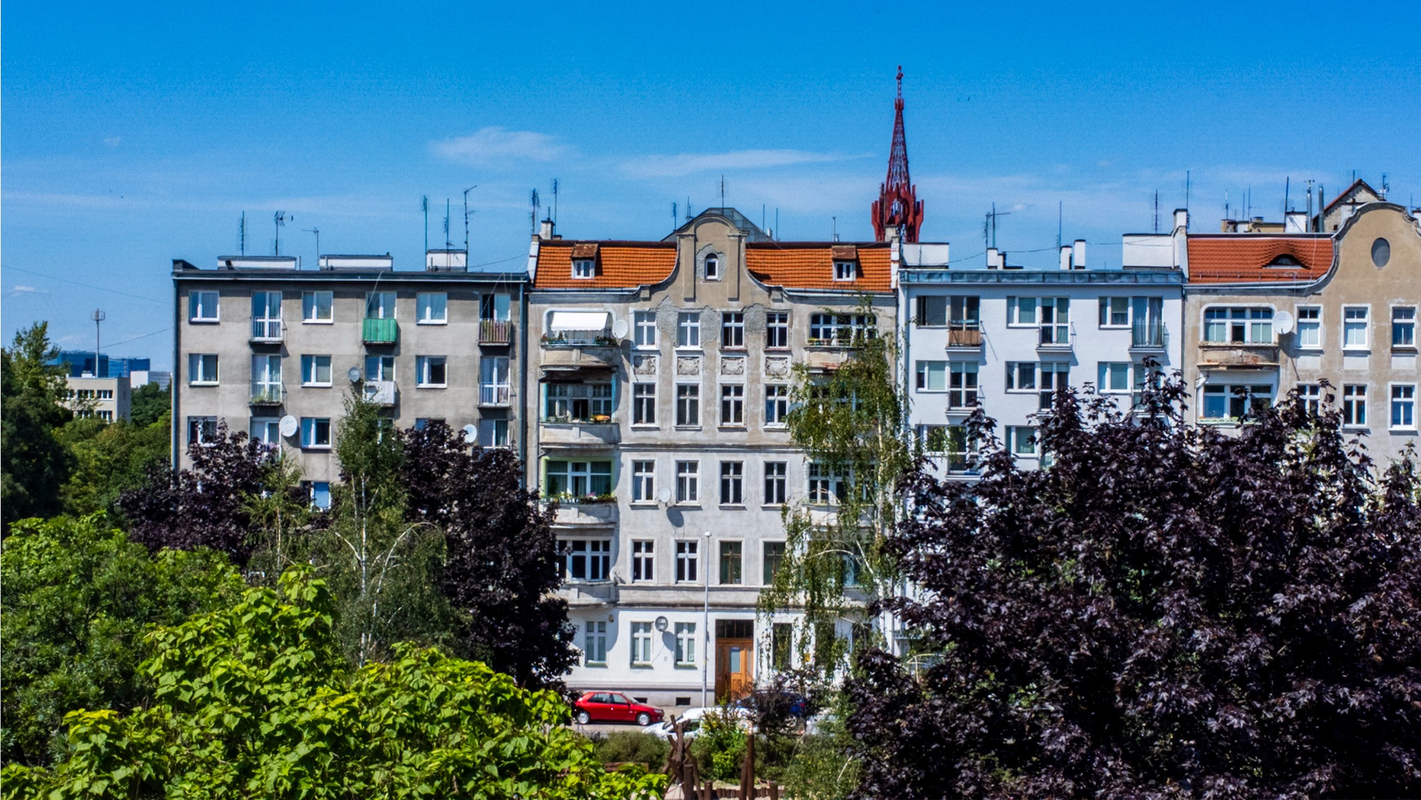Fasada kamienicy z czerwonym dachem i wieżą kościoła w tle, przesłonięta drzewami.