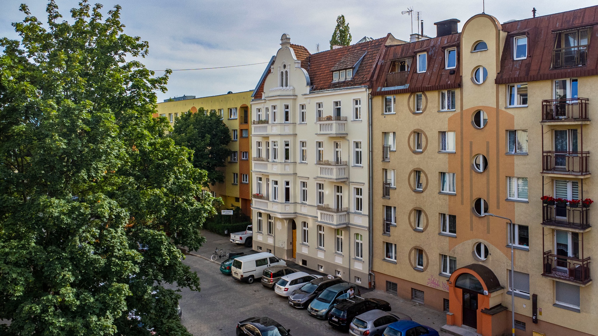 Widok z góry na ulicę z zaparkowanymi samochodami i fasady kamienic o zróżnicowanej architekturze i kolorystyce, z dużym drzewem po lewej stronie kadru.