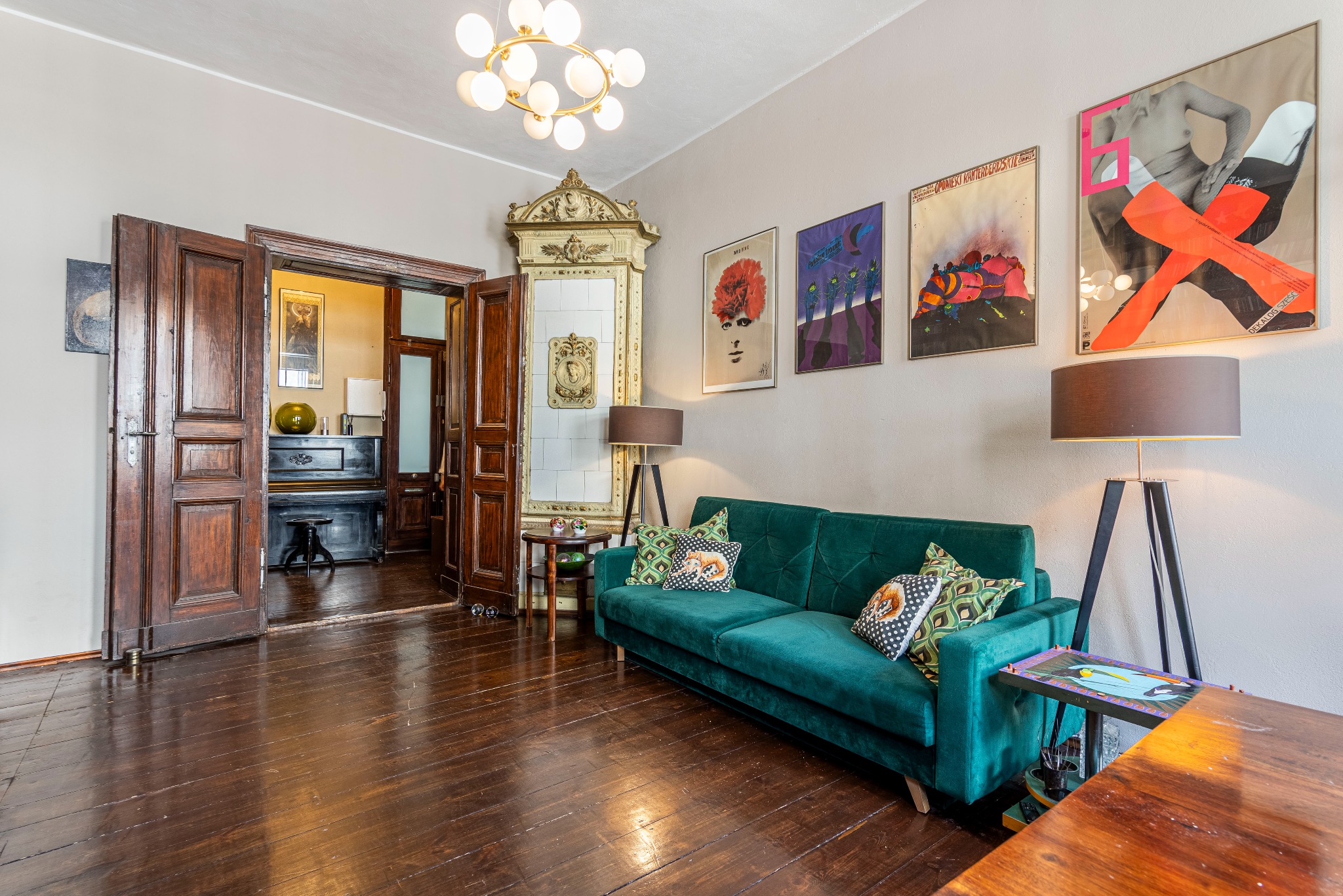 Wnętrze salonu z ciemną drewnianą podłogą, zieloną sofą, lampami podłogowymi, ozdobnym piecem kaflowym i plakatami na ścianie.