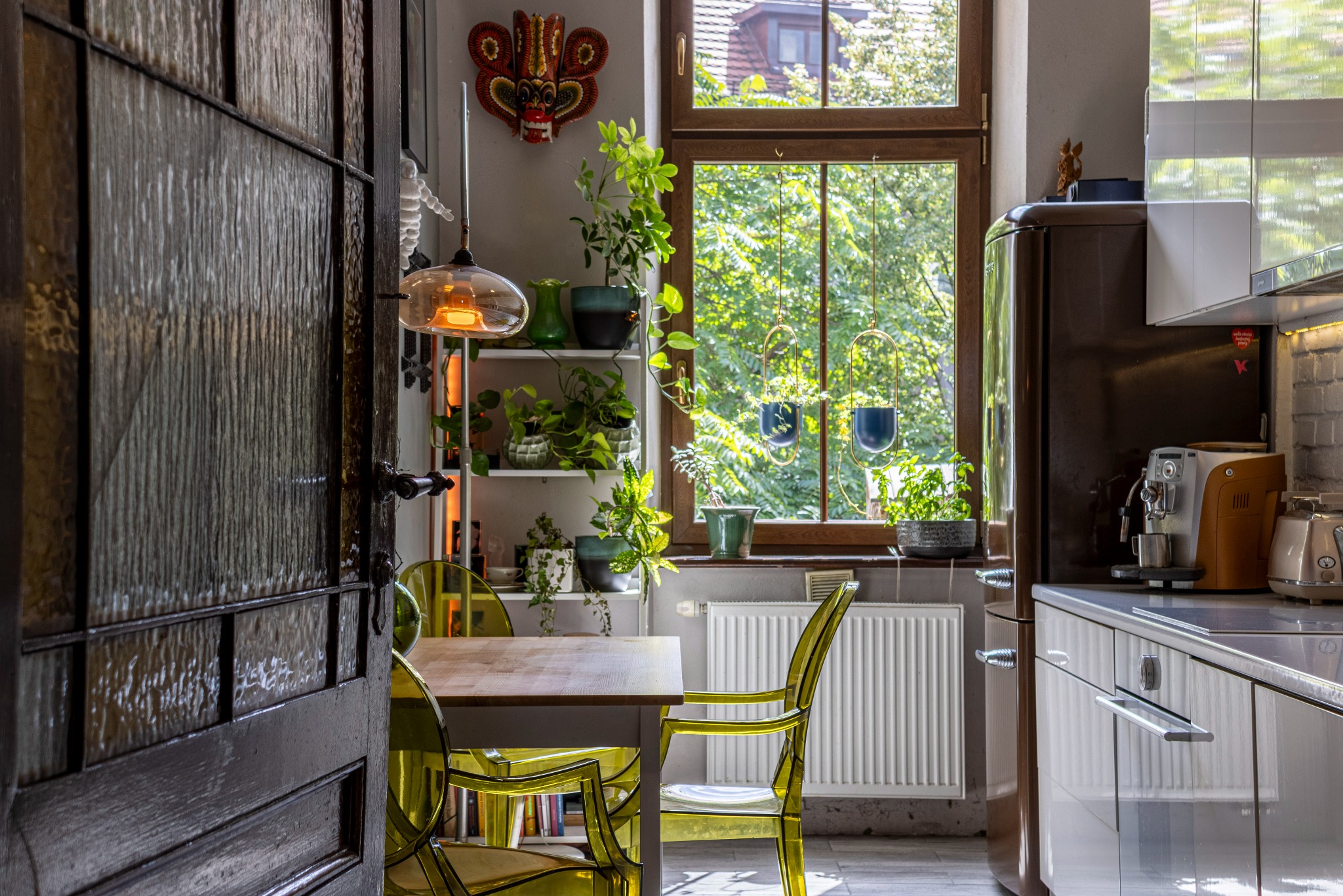 Jasna kuchnia z drewnianym stołem i żółtymi krzesłami z tworzywa, widok na okno z roślinami i dekoracyjną maską na ścianie, otwarta drewniana skrzydłowa drzwi.