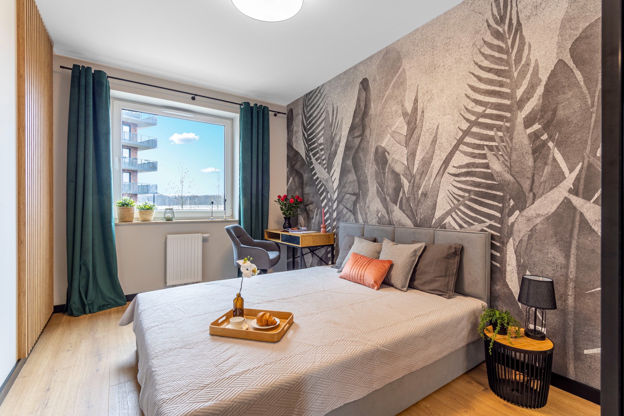 Jasna sypialnia z łóżkiem, stolikiem nocnym, biurkiem, oknem z widokiem na budynek i dekoracyjną tapetą z motywem liści.