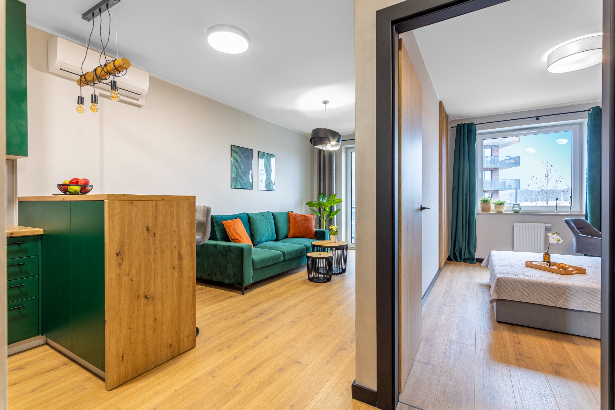 Jasne, nowocześnie urządzone wnętrze mieszkania z zieloną sofą, drewnianymi akcentami i widokiem na pokój z łóżkiem przez otwarte drzwi.