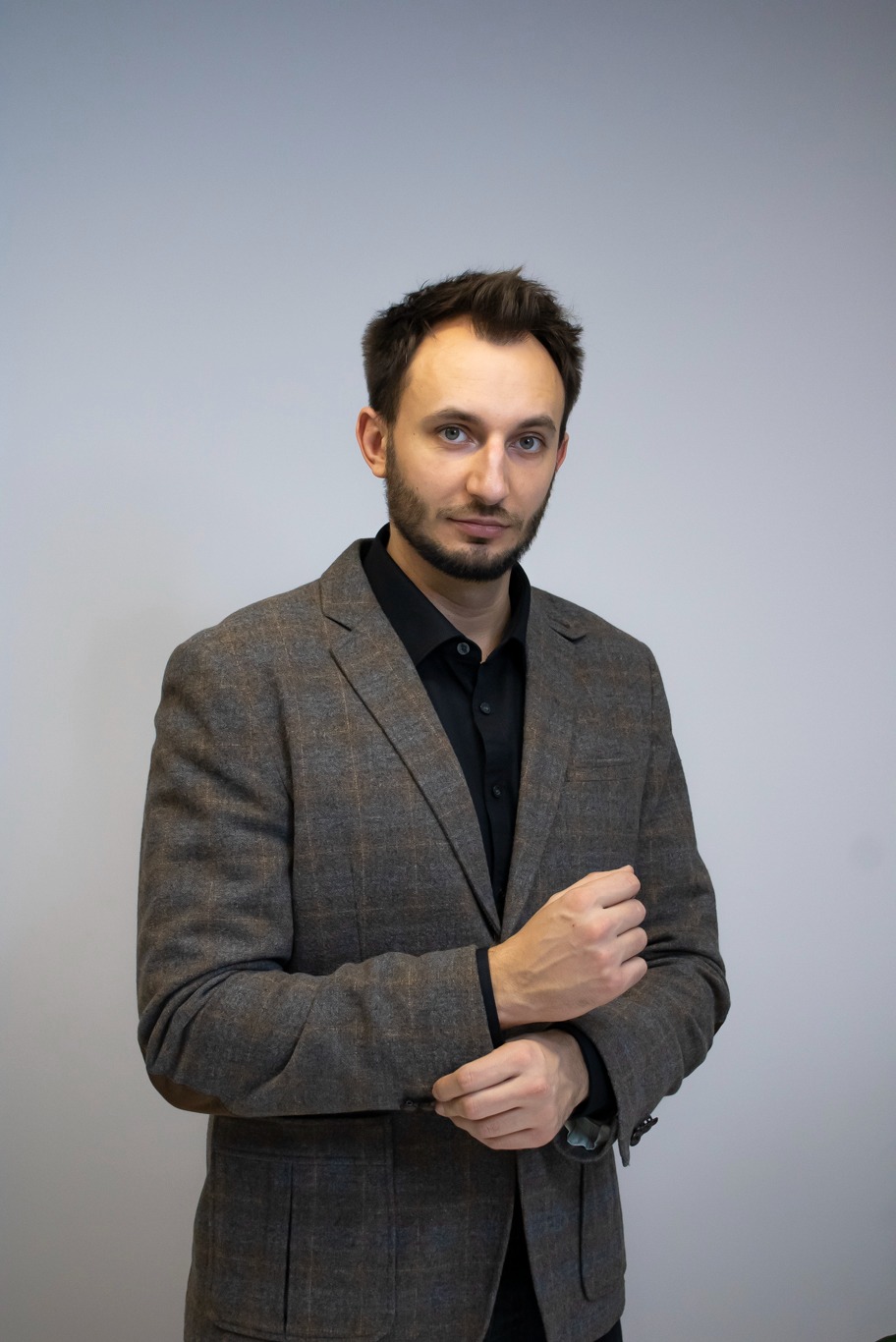 Portret mężczyzny w brązowej marynarce w kratę i czarnej koszuli, z zegarkiem na nadgarstku, na jasnoszarym tle.