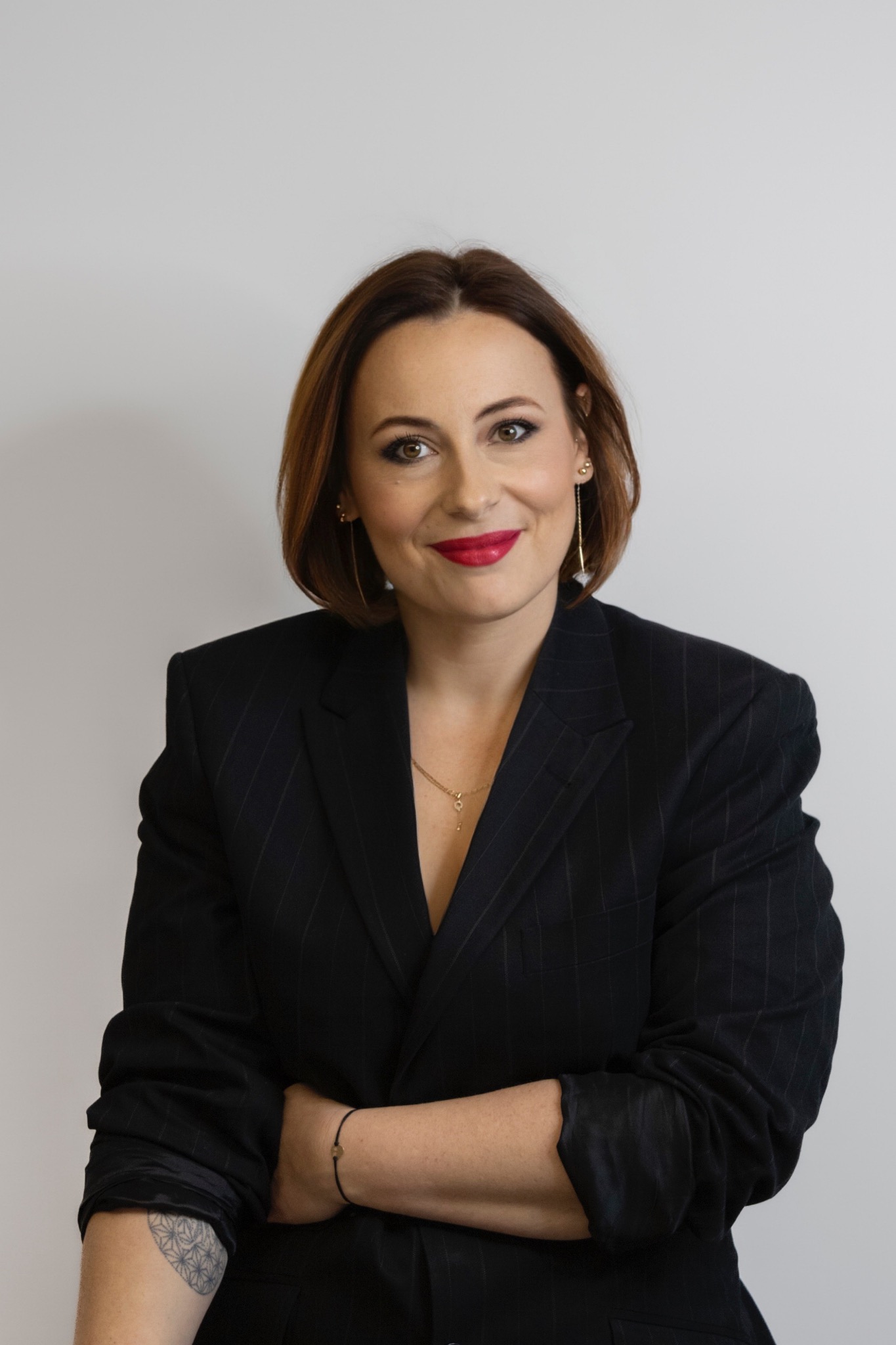 Portret uśmiechniętej kobiety w czarnym garniturze w prążki, z czerwoną szminką i złotą biżuterią, na jasnym tle, z widocznym tatuażem na ramieniu.