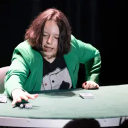 Młoda iluzjonistka w zielonej marynarce prezentuje sztuczkę z kartami i kostkami na zielonym stole, na tle czarnej kurtyny.