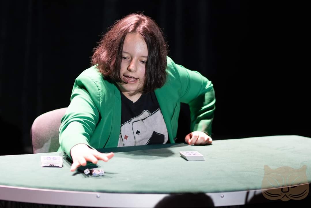 Młoda iluzjonistka w zielonej marynarce prezentuje sztuczkę z kartami i kostkami na zielonym stole, na tle czarnej kurtyny.