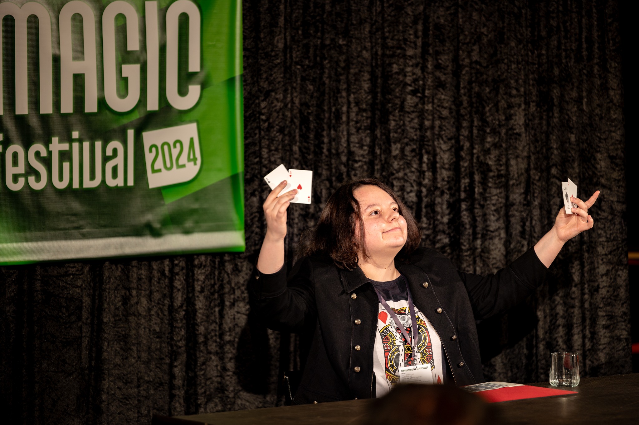 Kobieta w czarnej marynarce prezentuje karty podczas Magic Festival 2024, na stole szklanka i czerwona podkładka.