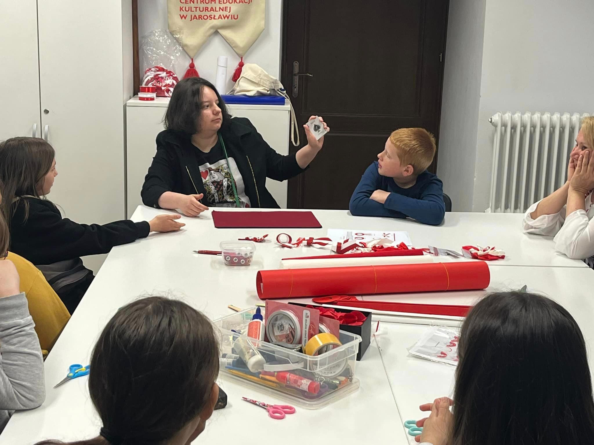 Kobieta w czarnej marynarce prezentuje kartę do gry (pik) grupie dzieci siedzących przy stole z materiałami plastycznymi, w tle widoczny napis 'Centrum Edukacji Kulturalnej w Jarosławiu'.