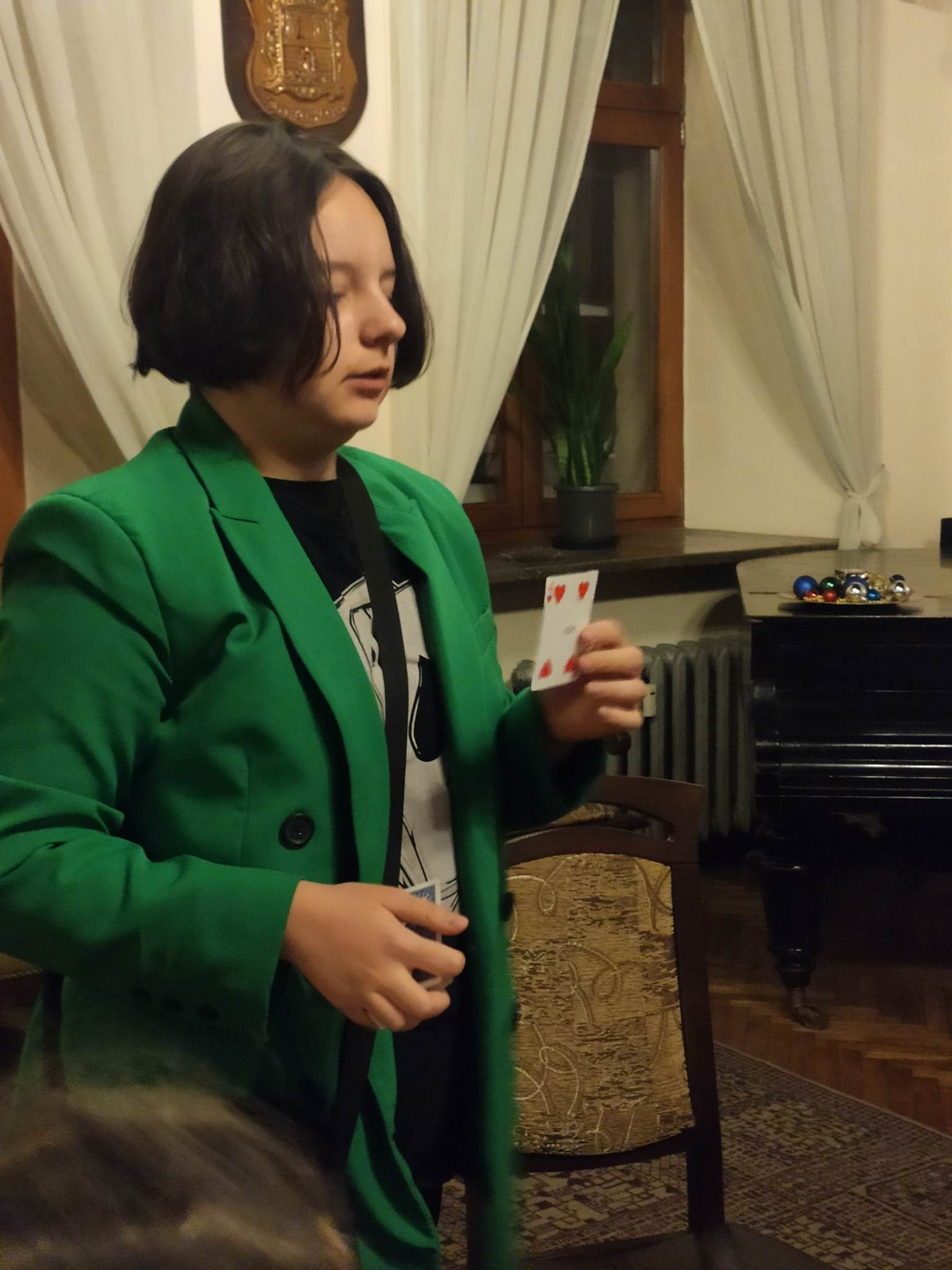 Osoba w zielonej marynarce trzymająca kartę do gry (czwórka kier) na tle pianina i dekoracji wnętrza.