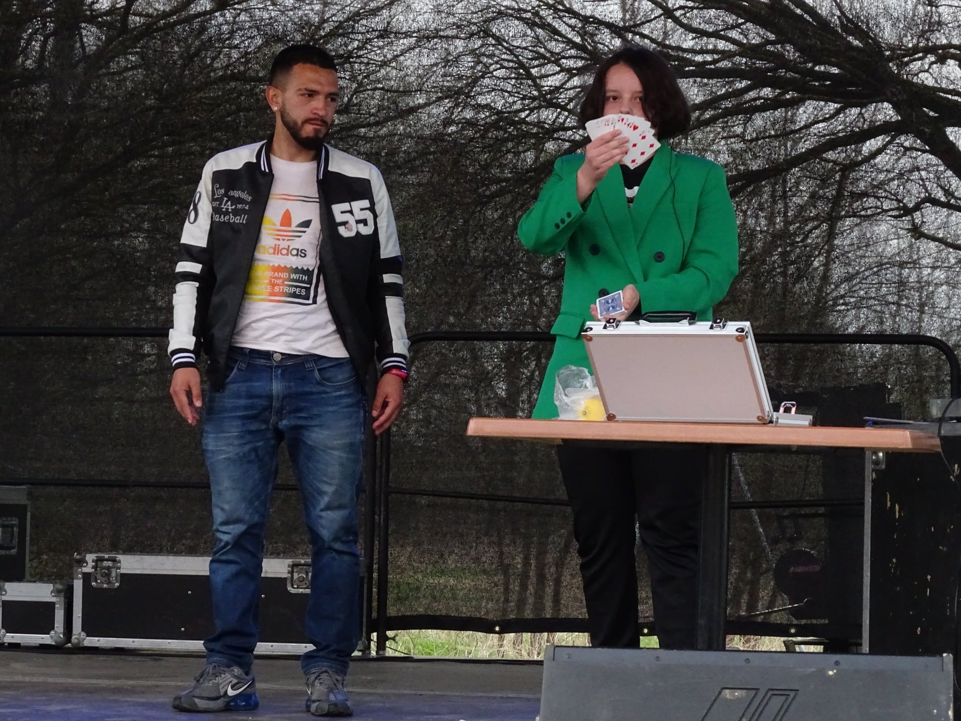 Młoda iluzjonistka w zielonym żakiecie prezentuje wachlarz kart pikowych, obok stoi mężczyzna w kurtce bejsbolówce, na stole srebrna walizka i rekwizyty.