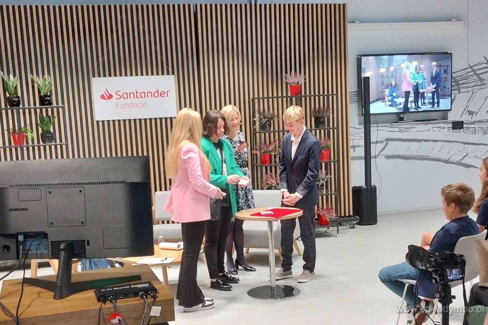 Młody iluzjonista w garniturze prezentuje trik z kartami na małym okrągłym stoliku, otoczony przez trzy kobiety w eleganckich strojach, w tle logo Santander Fundacja i monitor pokazujący tę samą...