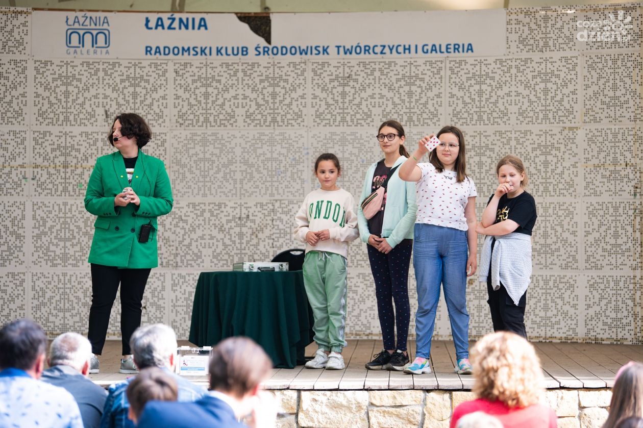 Iluzjonistka w zielonej marynarce prezentuje sztuczkę karcianą z udziałem młodych dziewcząt na scenie plenerowej, publiczność obserwuje występ.