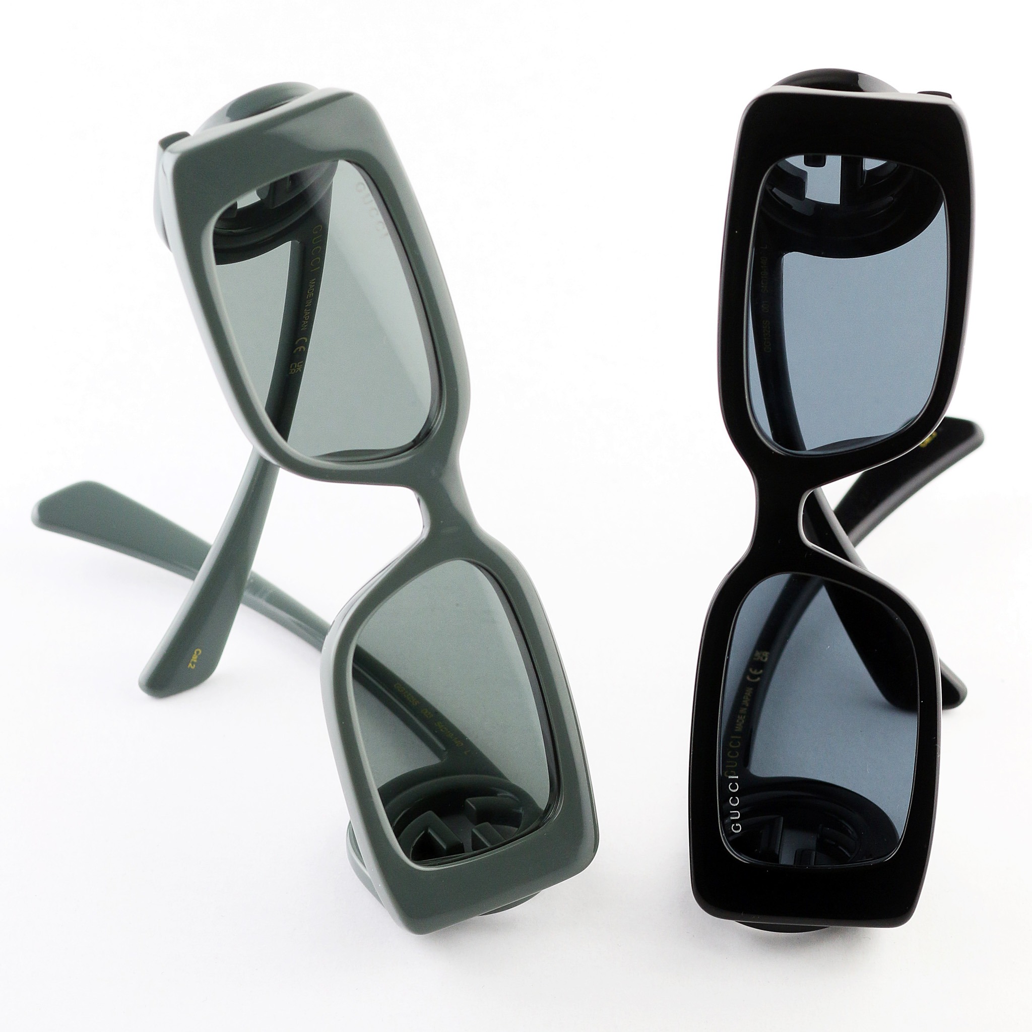 Dwie pary okularów przeciwsłonecznych marki Gucci: jedna w kolorze oliwkowym, druga czarna, ustawione pionowo na białym tle, eksponujące logo marki.
