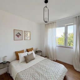 HOME INVEST PARTNERS SPÓŁKA Z OGRANICZONĄ ODPOWIEDZIALNOŚCIĄ - Jasna sypialnia z łóżkiem z pikowaną narzutą w drobne kwiatki, drewnianą podłogą i minimalistyczną lampą wiszącą o owalnym kształcie.