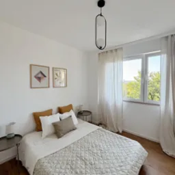 Jasna sypialnia z łóżkiem z pikowaną narzutą w drobne kwiatki, drewnianą podłogą i minimalistyczną lampą wiszącą o owalnym kształcie.